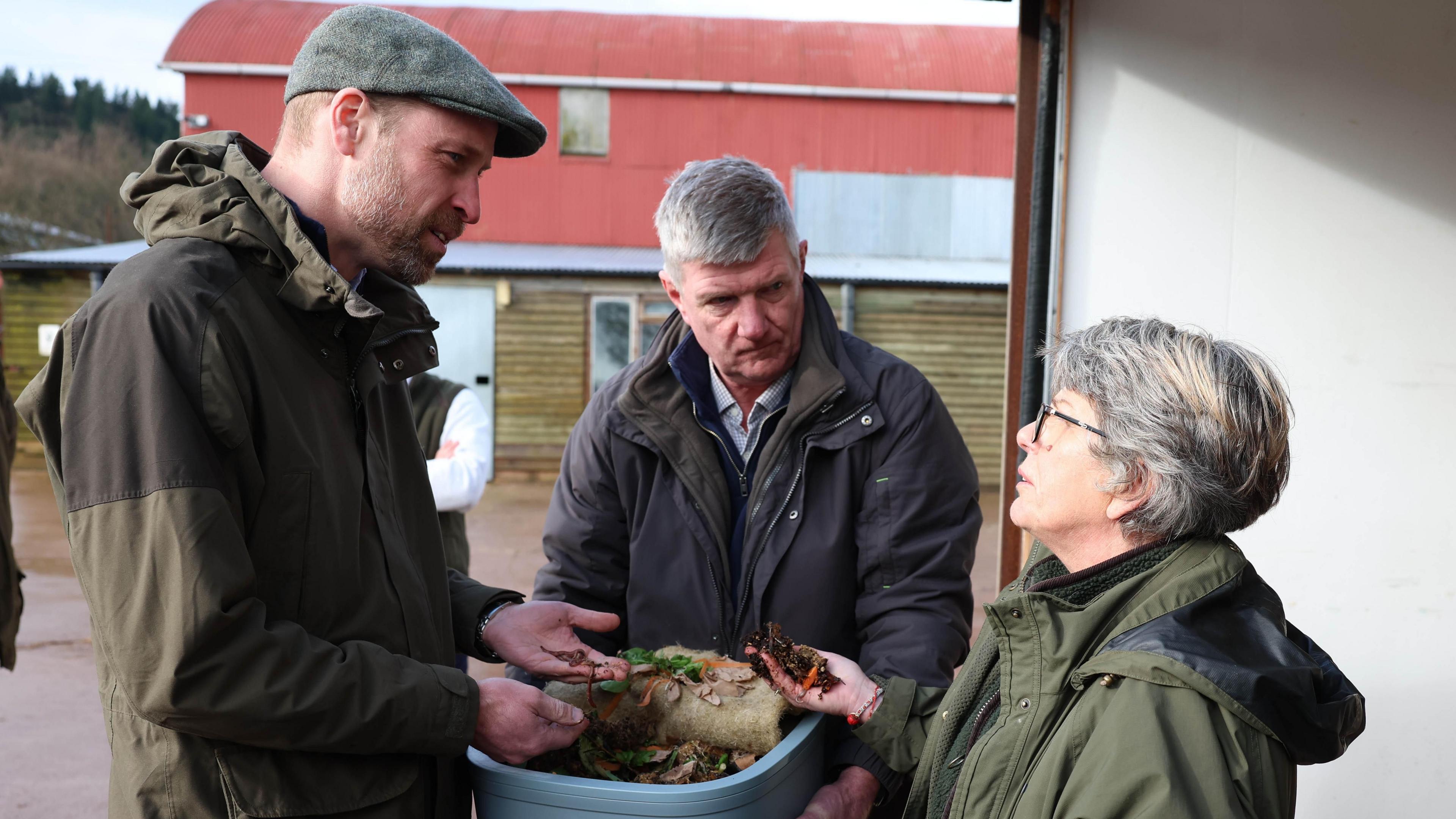 The Prince of Wales stands with his hands over a bucket filled with food waste. In his hands are some worms. He is speaking to a man and a woman, who is also holding composting material.