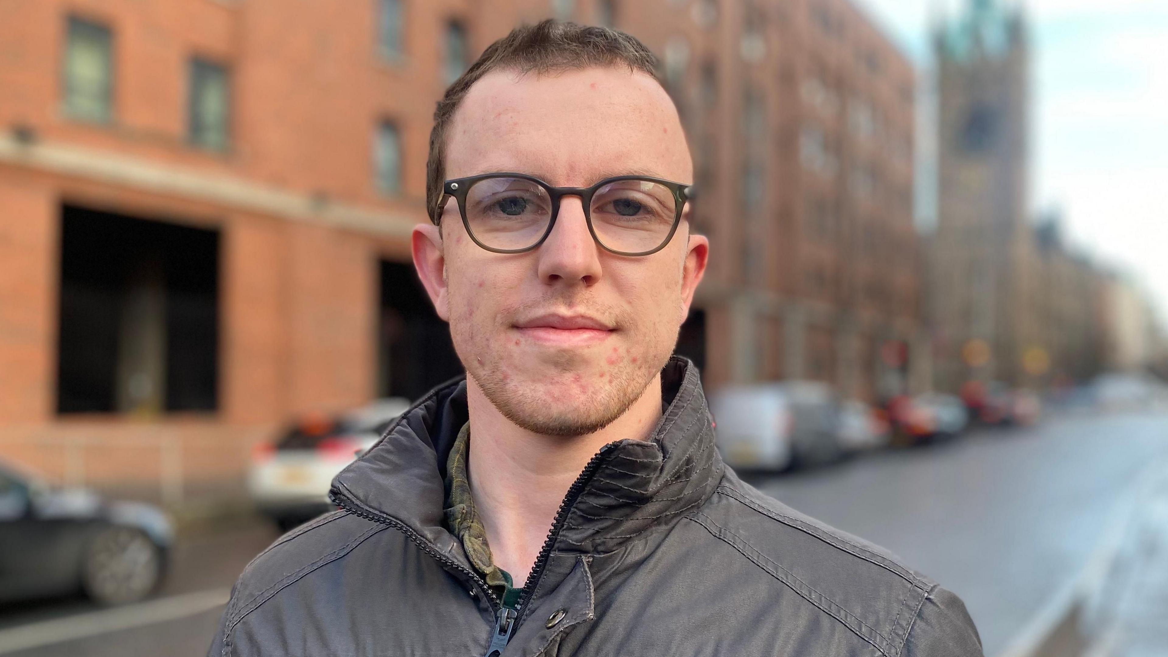 Matthew Green wears glasses and a brown coat. He has brown hair. There are cars and buildings on the street behind him.