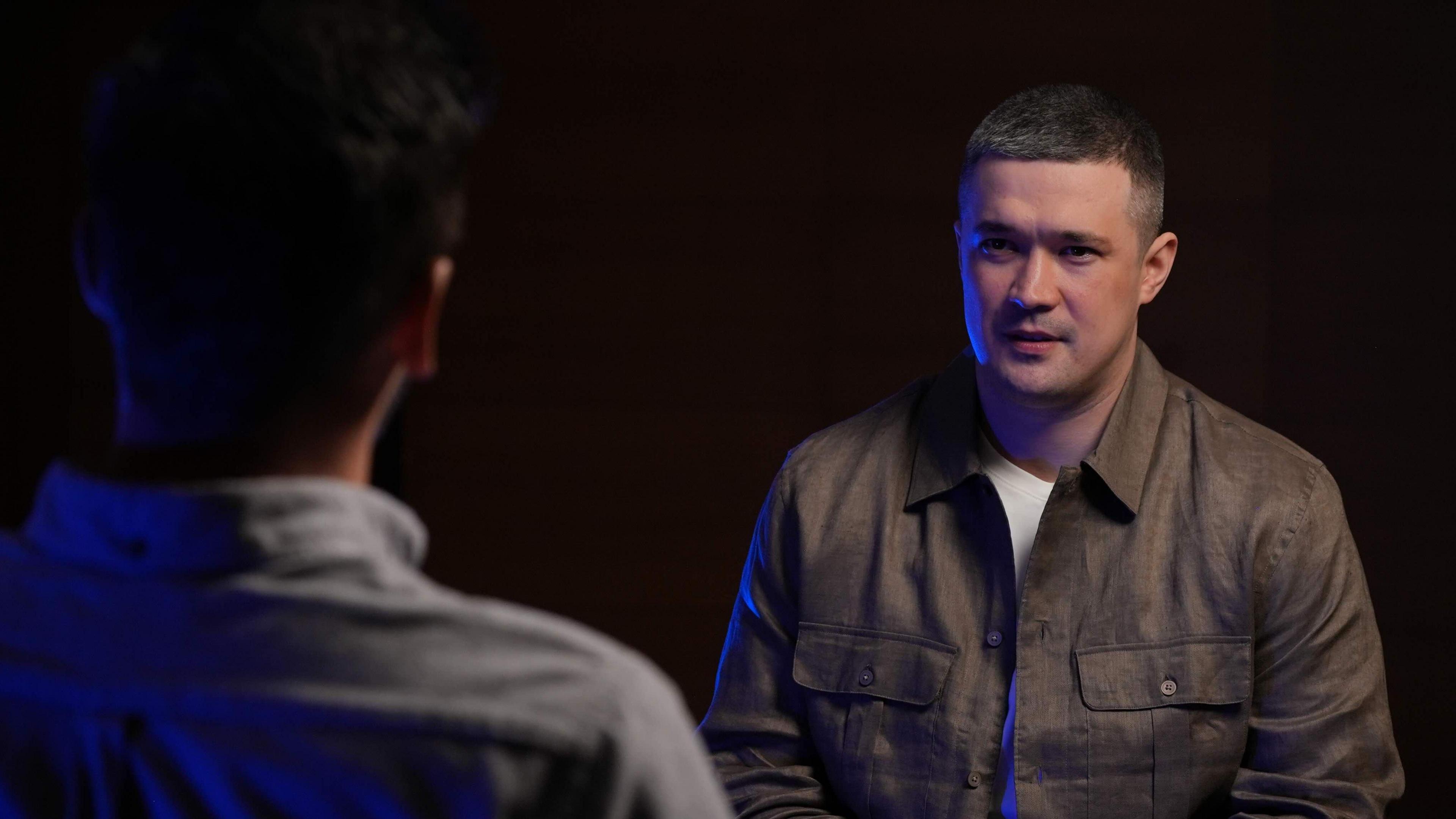 A man dressed in a white T-shirt and brown collared shirt in front of a black background sits talking to someone with their back to the camera.