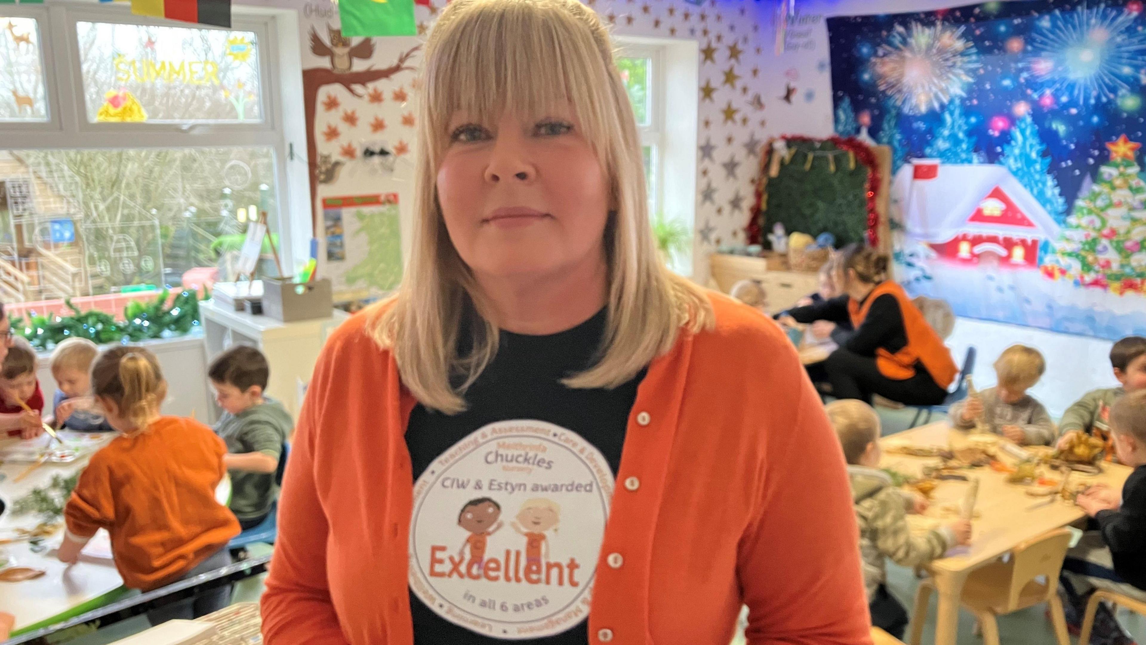 Lisa Owen, wearing a Chuckles nursery T-shirt, showing its "excellent" rating, in the nursery while children work at tables behind, with Christmas pictures on the wall