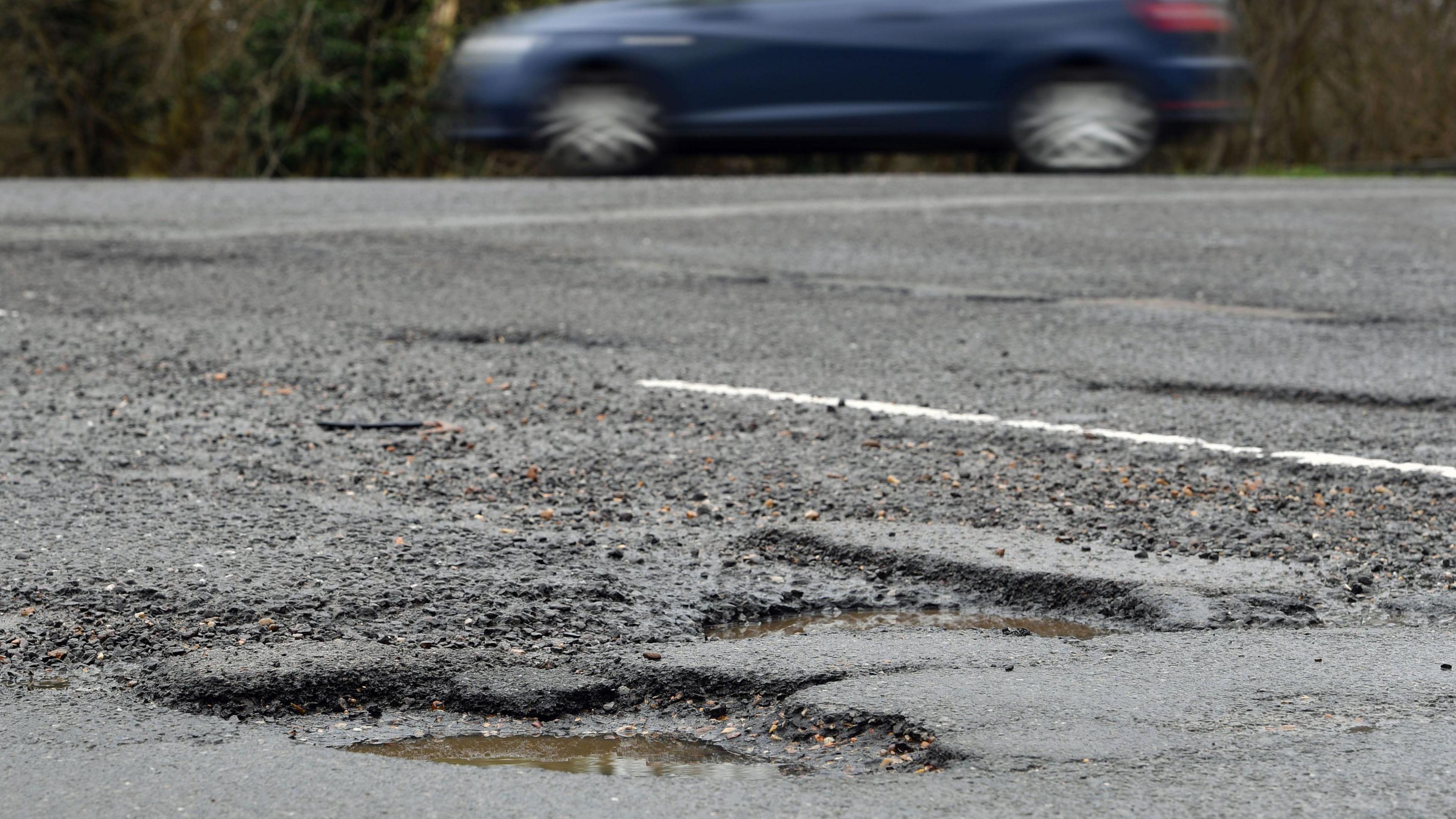 Blurred blue car passing potholes, which are filled with rainwater. The shot focuses on the potholes