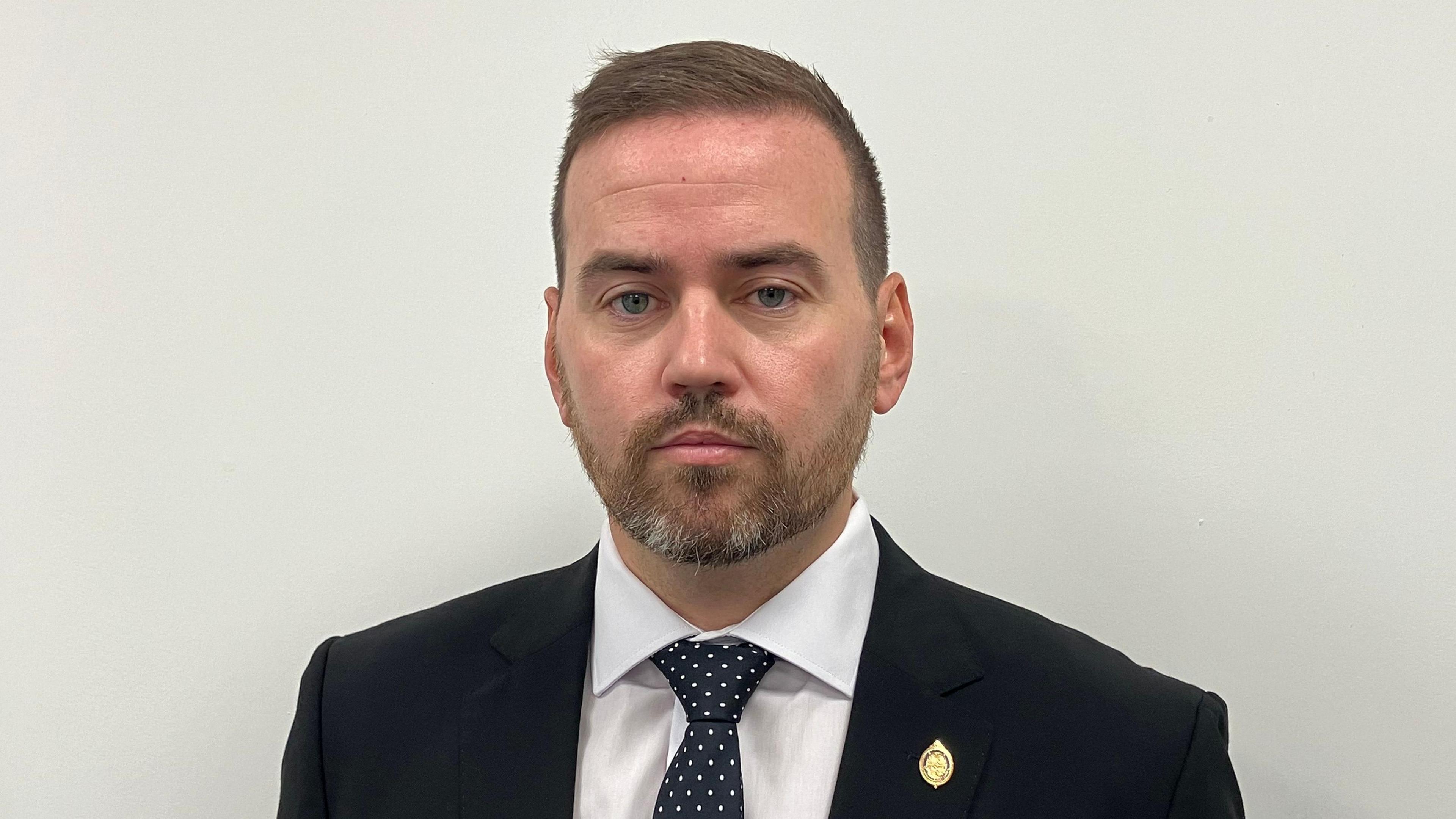 Gavin Callaghan appearing with a beard and short hair. He is wearing a black or navy blue suit, a white shirt and a navy blue tie with white spots on it. There is a cream or off-white painted wall behind him.