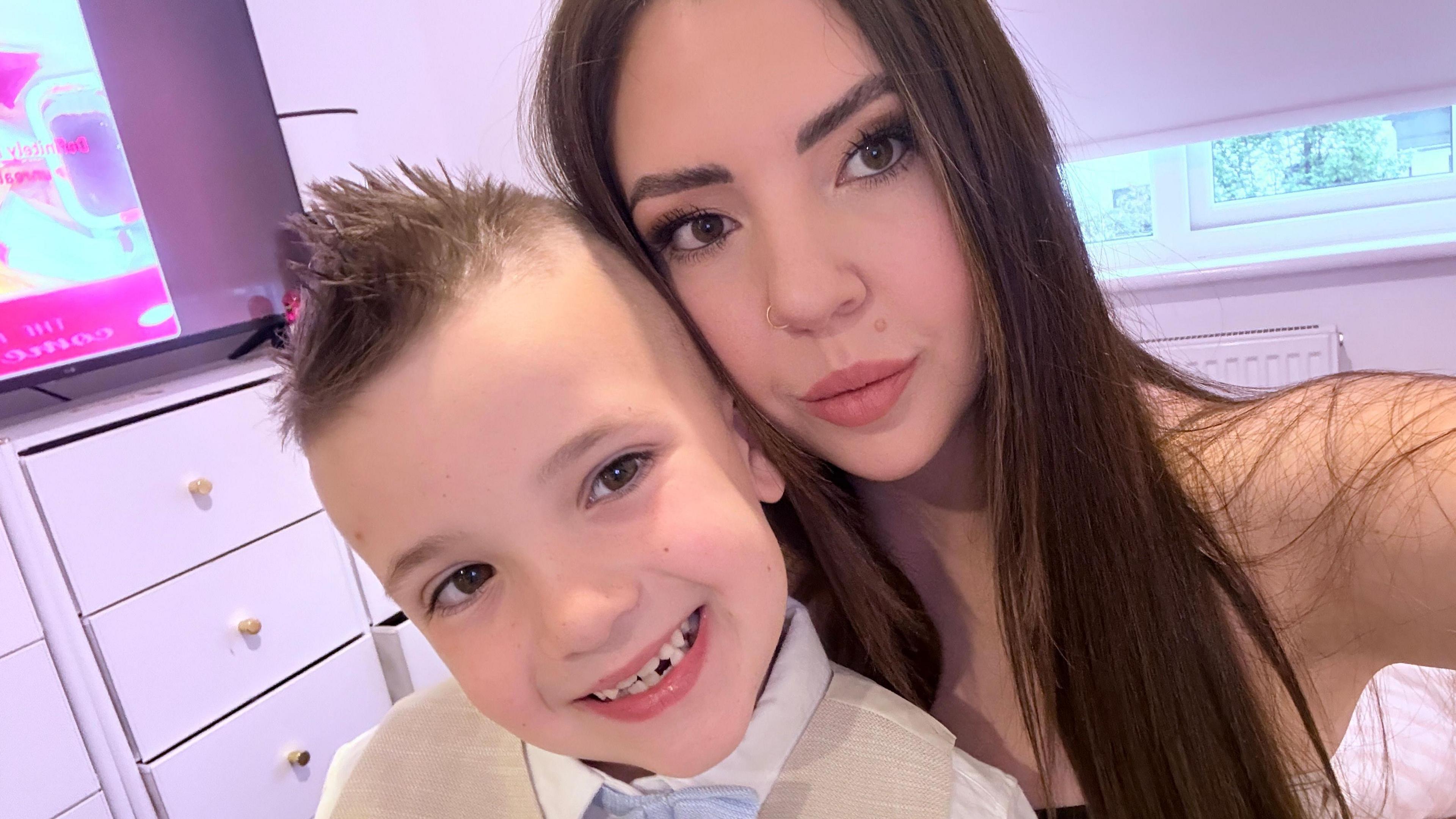 a boy with a smart waistcoast and bow tie smiles as he is hugged by his mum who has long brown hair and is pouting