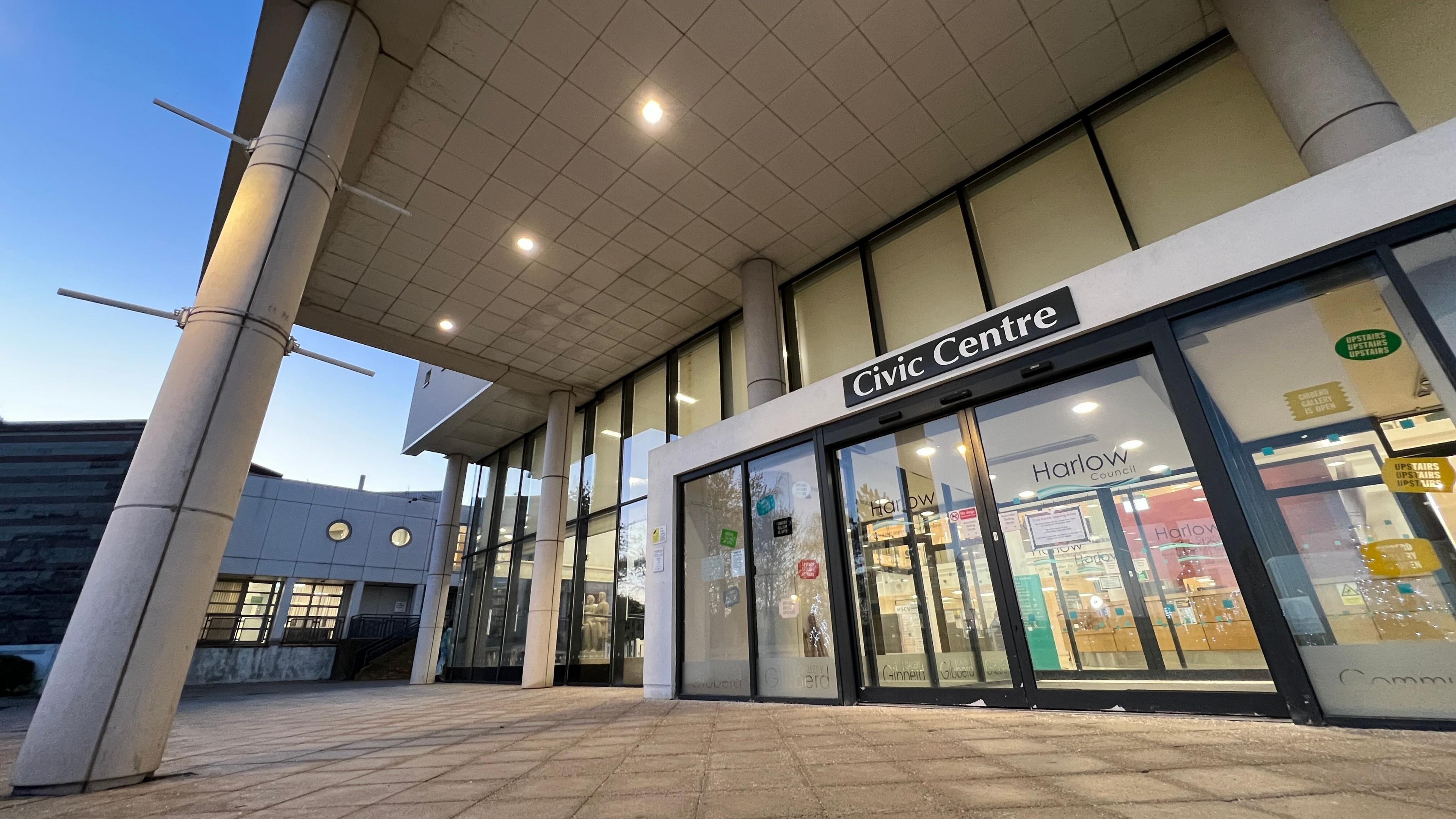 Harlow Civic Centre's main entrance, which contains the offices for Harlow Council. The photo is taken from outside the front doors. The inside is illuminated with lights. It is dusk outside, with blue skies above.