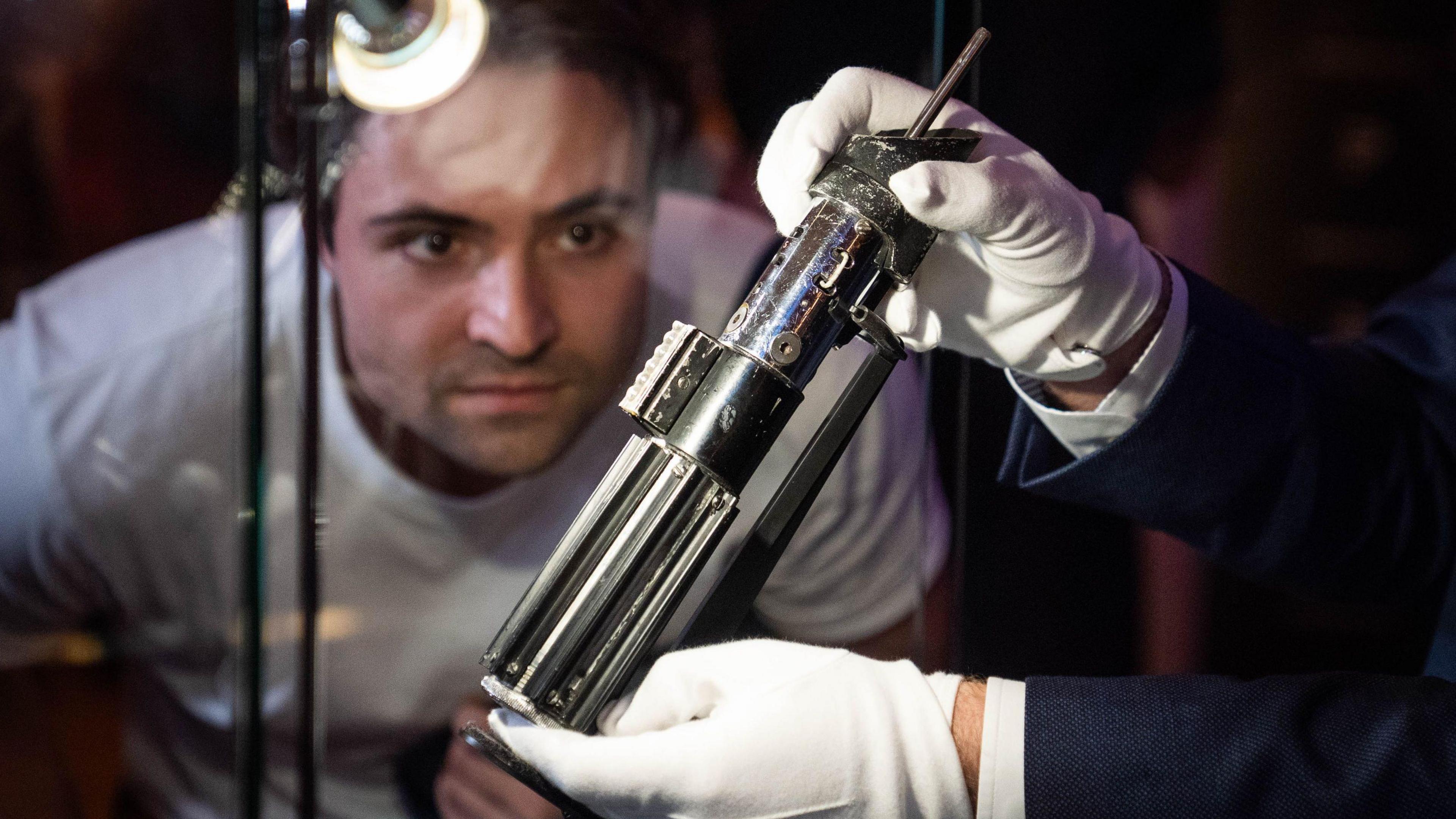 A man looks through the glass at the base of a silver lightsaber, held by someone using white gloves.
