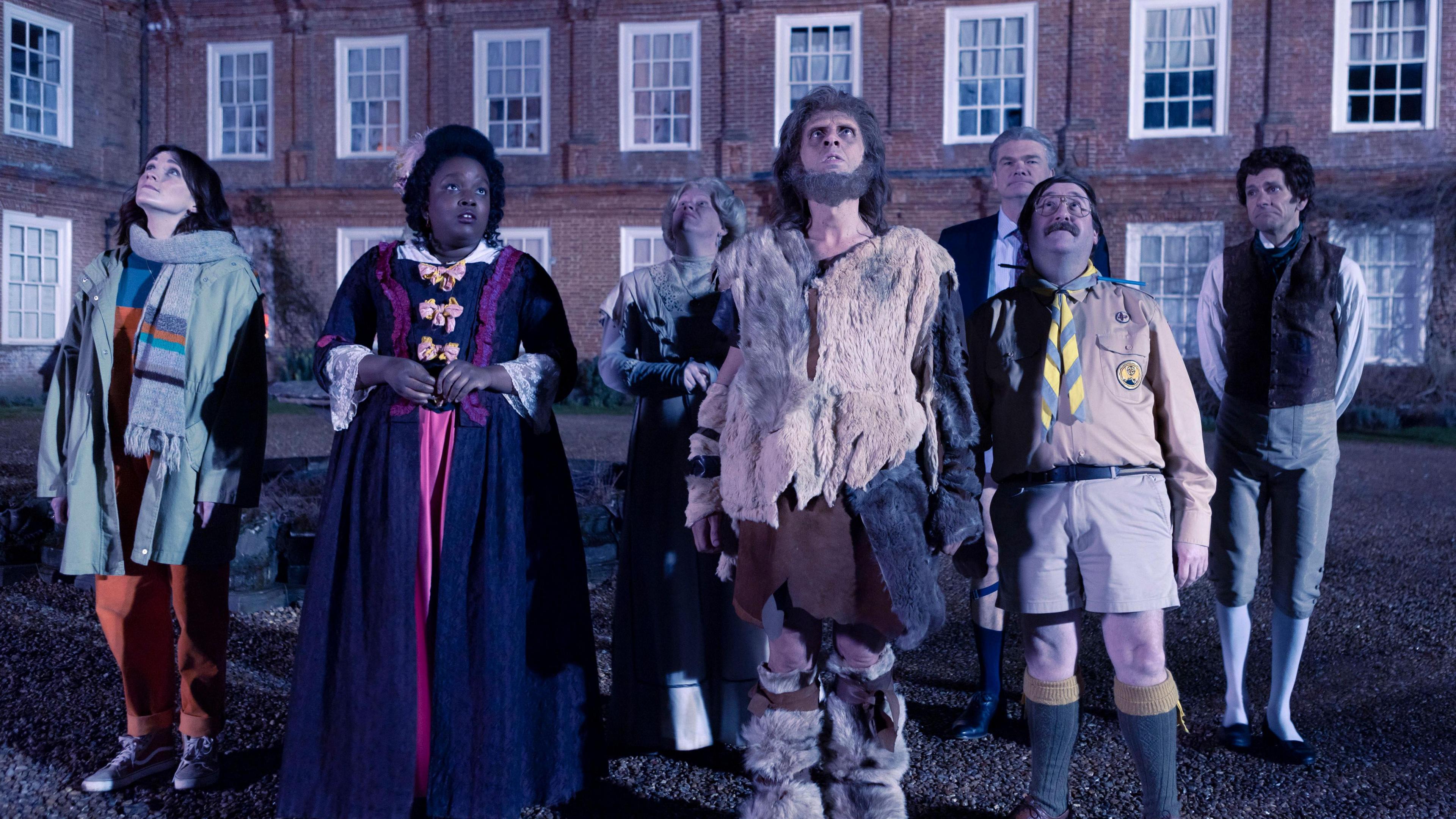 A screenshot of the TV show Ghosts - seven of the characters are standing outside the house looking up at the sky. One woman on the left is wearing modern clothes - orange dungarees, a Khaki-coloured coat and scarf. The other characters are in historic costumes.