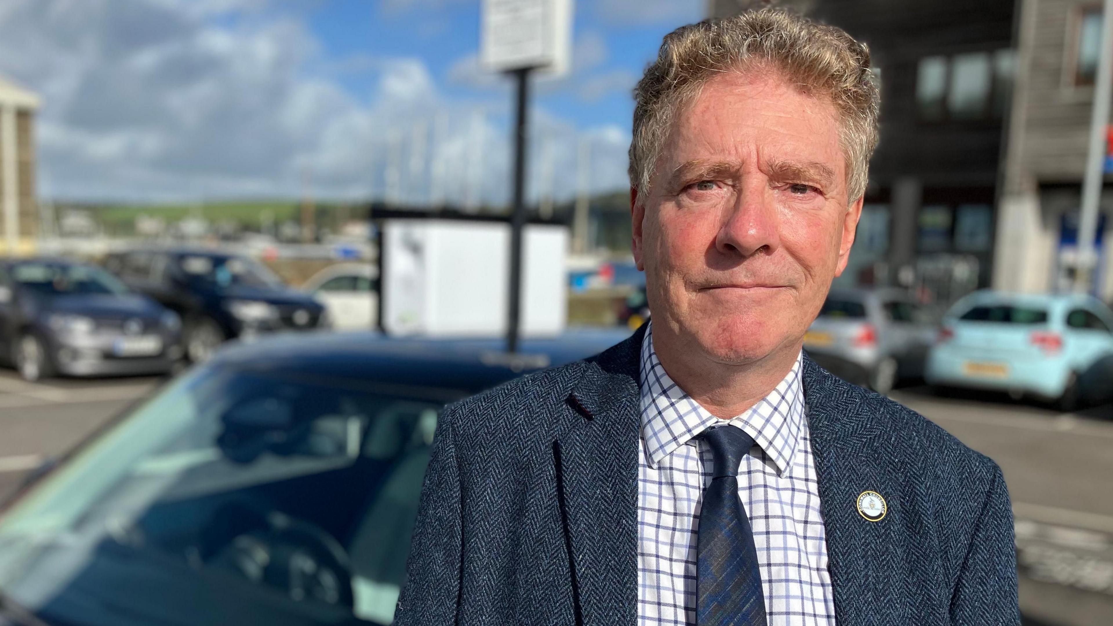 A man stands in a car park in front of an electric car and an electric car charger. He is wearing a blue jacket. 