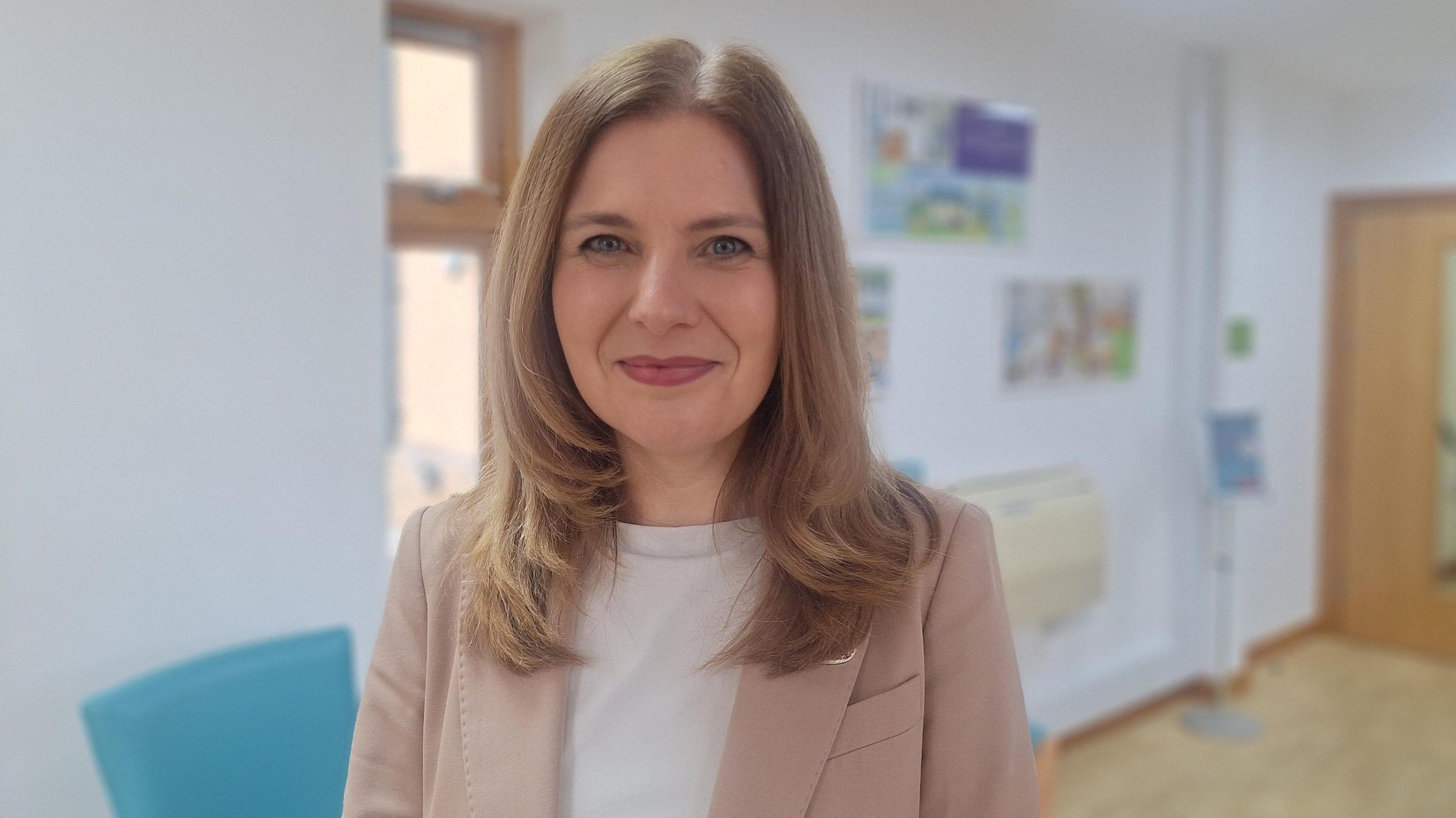 A woman stood in a corridor in a hospice. The background is blurred and she is in focus. She has shoulder-length blonde/brown hair and smiles at the camera. She has pink lipstick on and wears a beige jacket over a white t-shirt. It is a head and shoulders shot. 