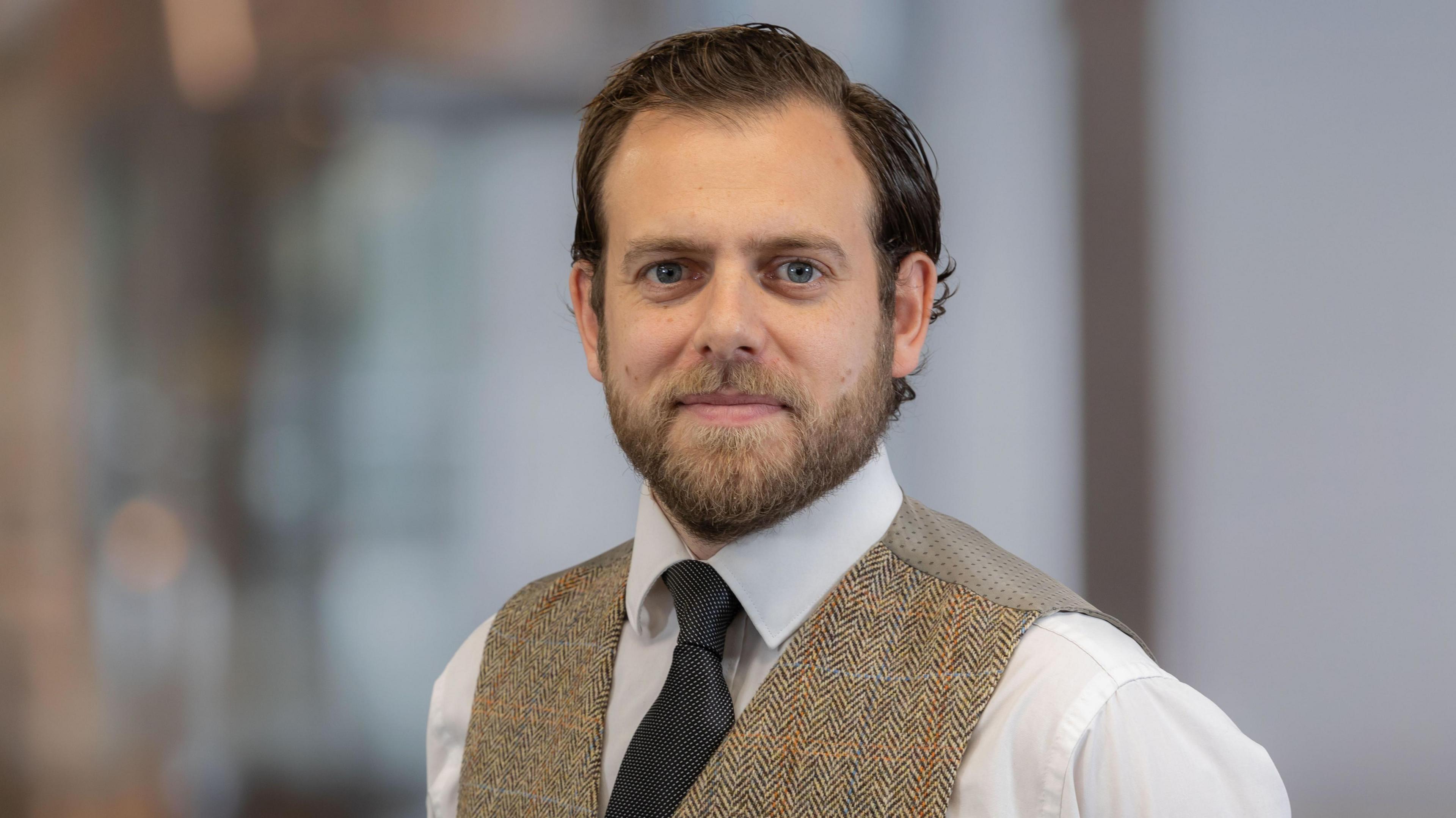 Stuart Leslie, a man with brown hair and a brown beard. He is wearing a white shirt, dark tie and a beige waistcoat. He is looking at the camera. The background is blurry.