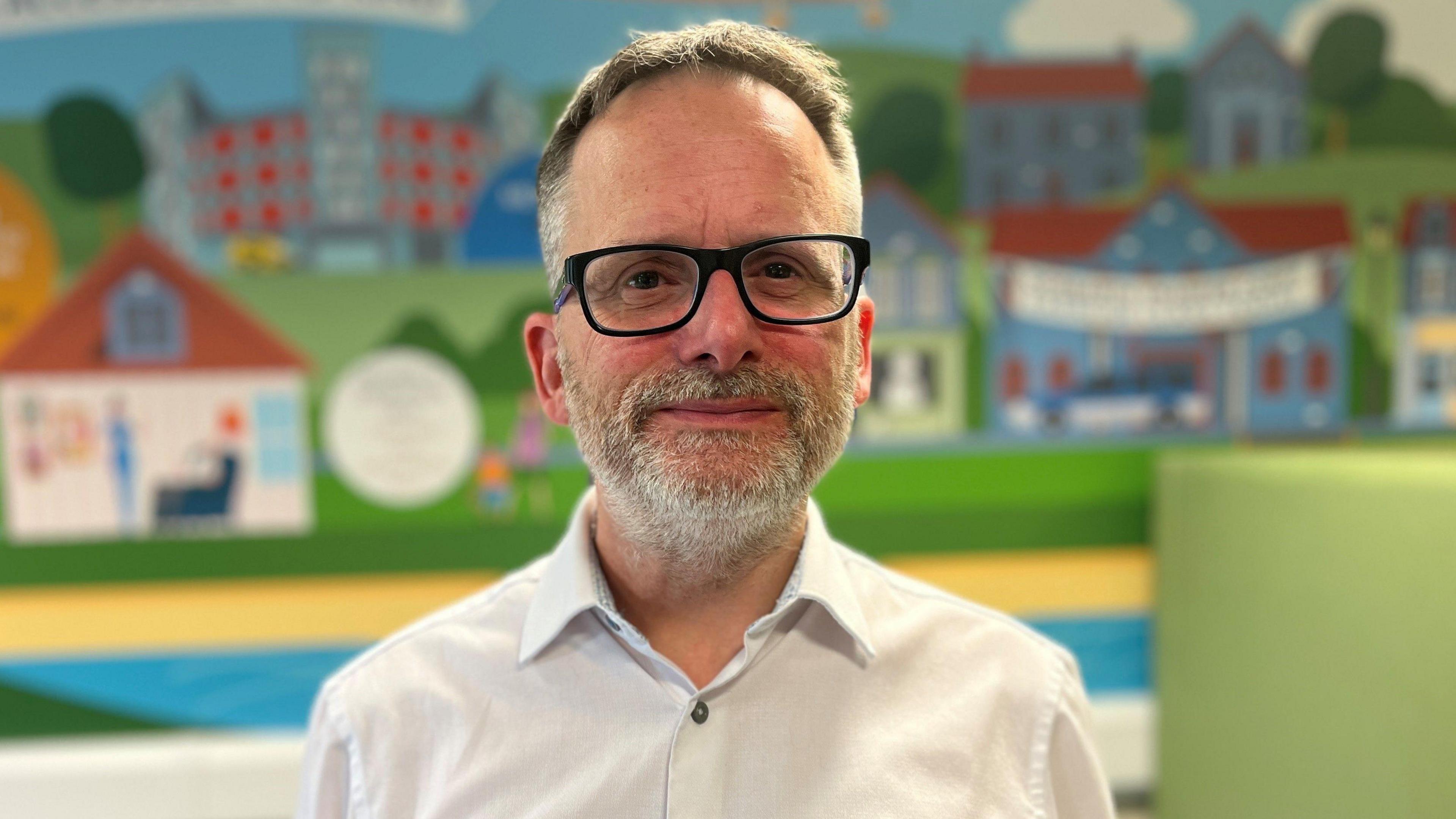 Phil Kloer looks at the camera. He has short grey hair and a short grey beard. He is wearing glasses and a white shirt and is standing in front of a mural with houses, the background is slightly out of focus.