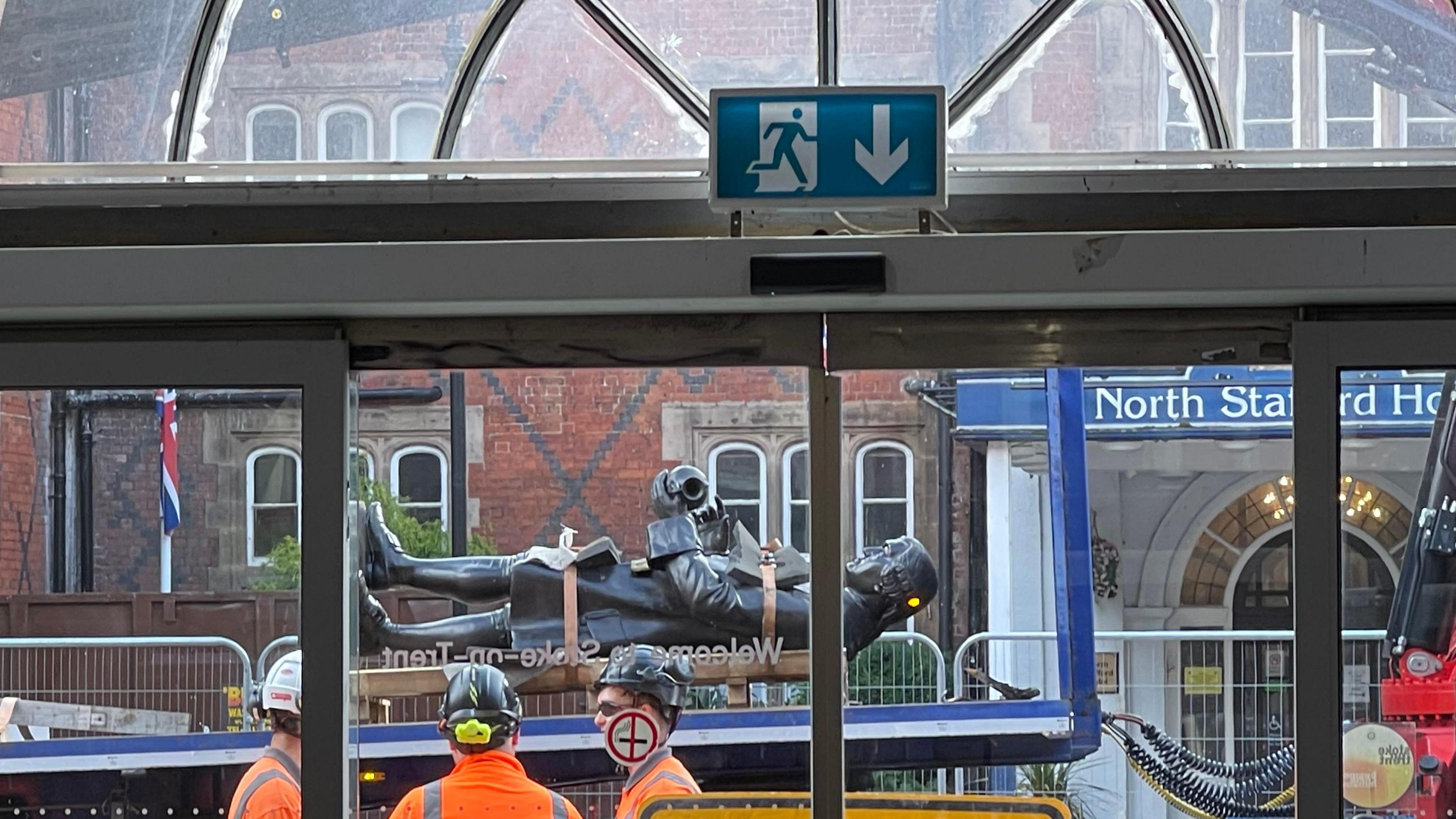A statue of potter Josiah Wedgwood, lay on its back, is being transported past the frontage of a railway station. It is viewed through a set of glass windows.