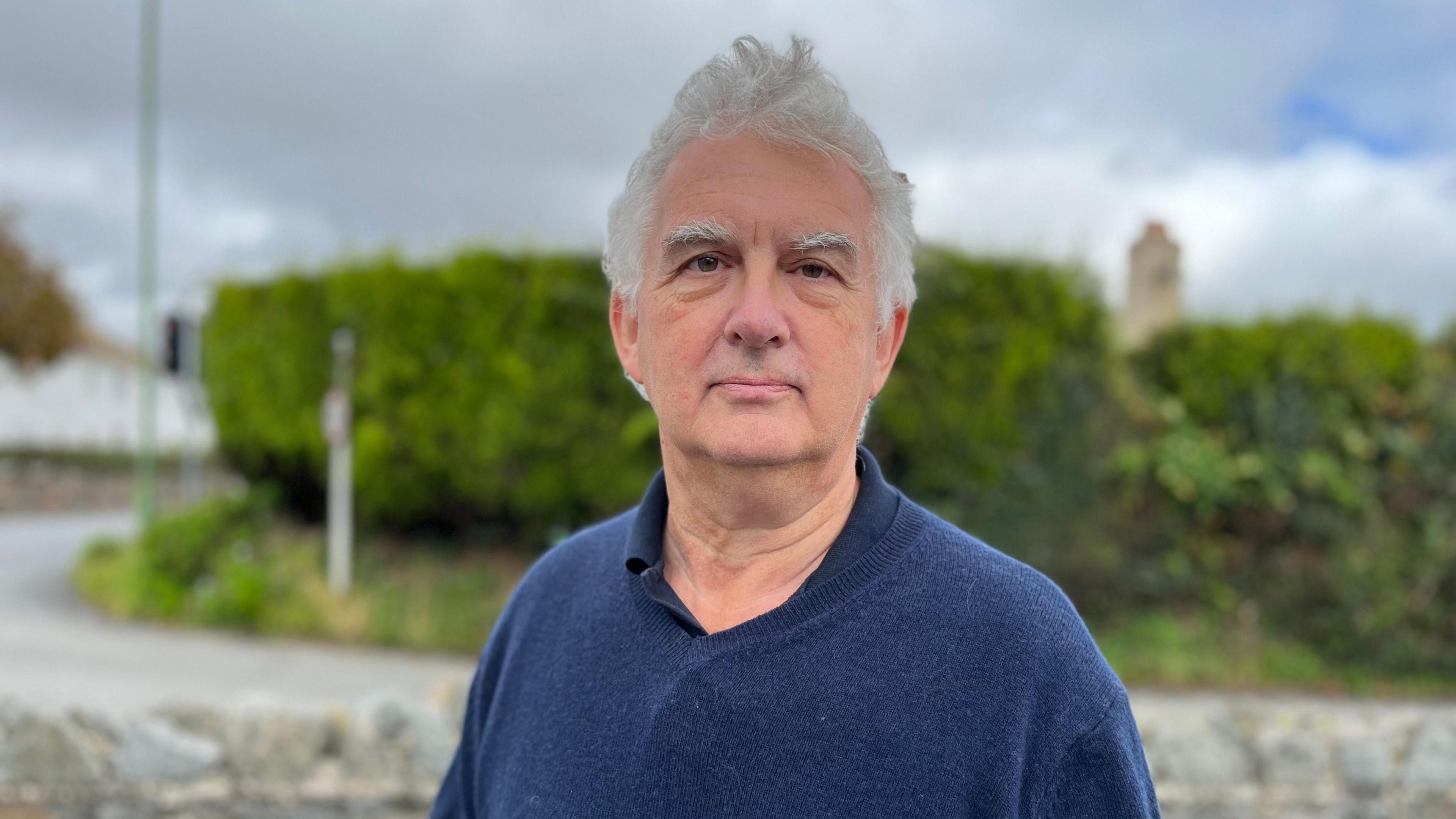 A man in a blue woollen jumper stands in front of a small granite wall and a neat hedge. He has bushy eyebrows and white, whispy hair.