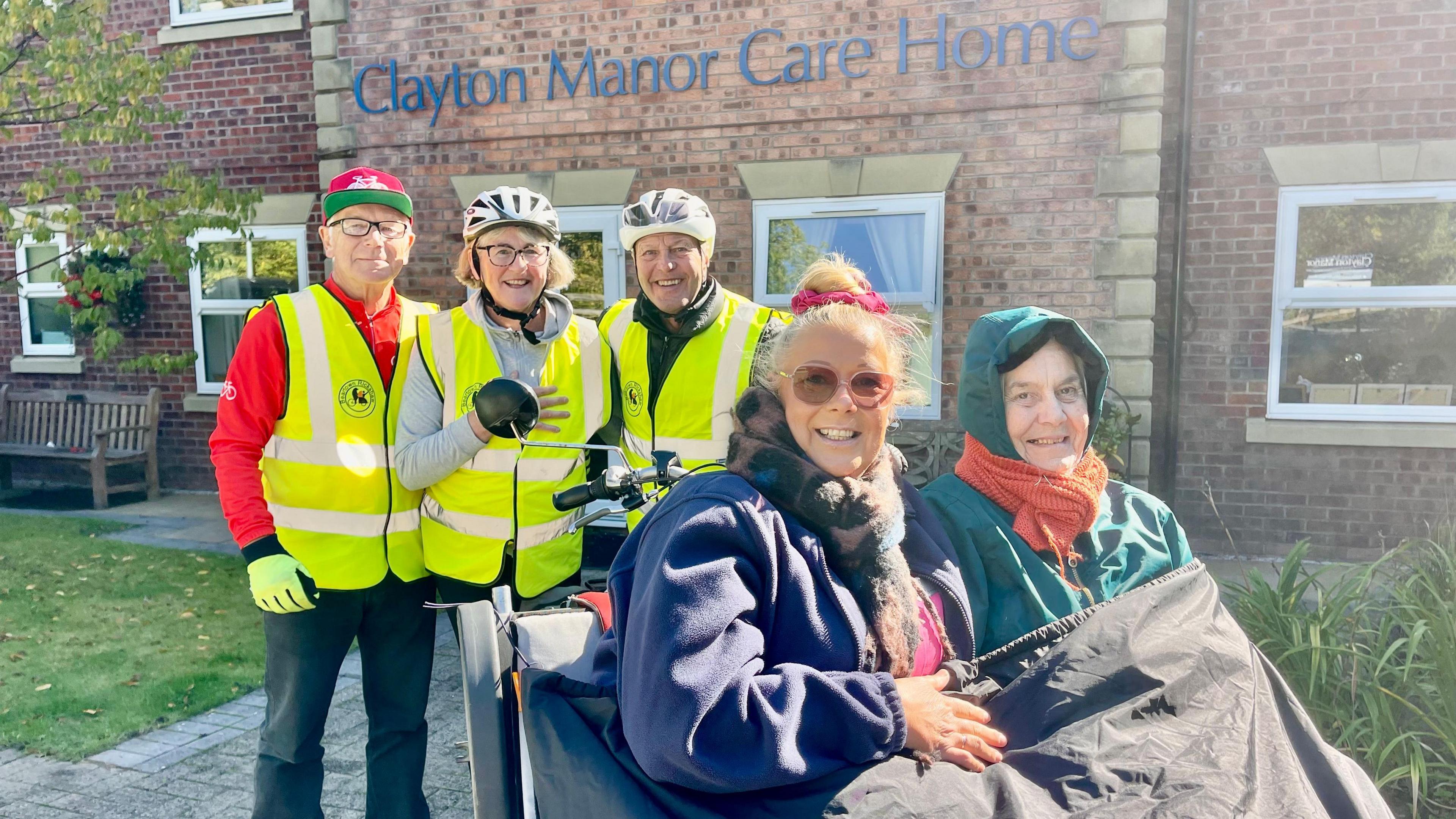 Two women sit on the front of a rickshaw. Behind them are two men and a woman. One man is wearing a white helmet, hi vis vest, black coat and black gloves. To the left of him is a woman also in a white helmet, hi vis vest, grey hoodie, glasses and has short blonde hair. To the far left is a man in a red baseball cap, glasses, red fleece, hi vis vest and black trousers. The elderly woman in the right seat is wearing a green coat with the hood up and an orange scarf. The woman in the right seat has blonde hair tied in a pink scrunchie, sunglasses, a dark scarf and a navy fleece. They are both covered by a black waterproof blanket wrapped over their legs. They are stood outside a building that has the words 'Clayton Manor Care Home' written in blue lettering on its wall. There are plants, a bench, paved area and grassy areas outside the care home.