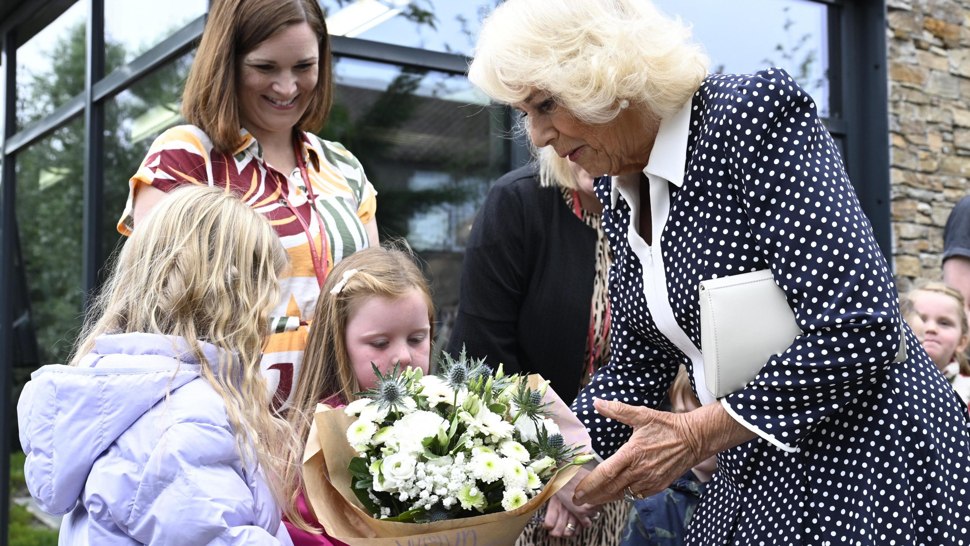 Queen Camilla wearing a black and white polka dot dress with a white collar received a bouquet of white flowers from two young girls. A woman standing behind the girls looks at them smiling.