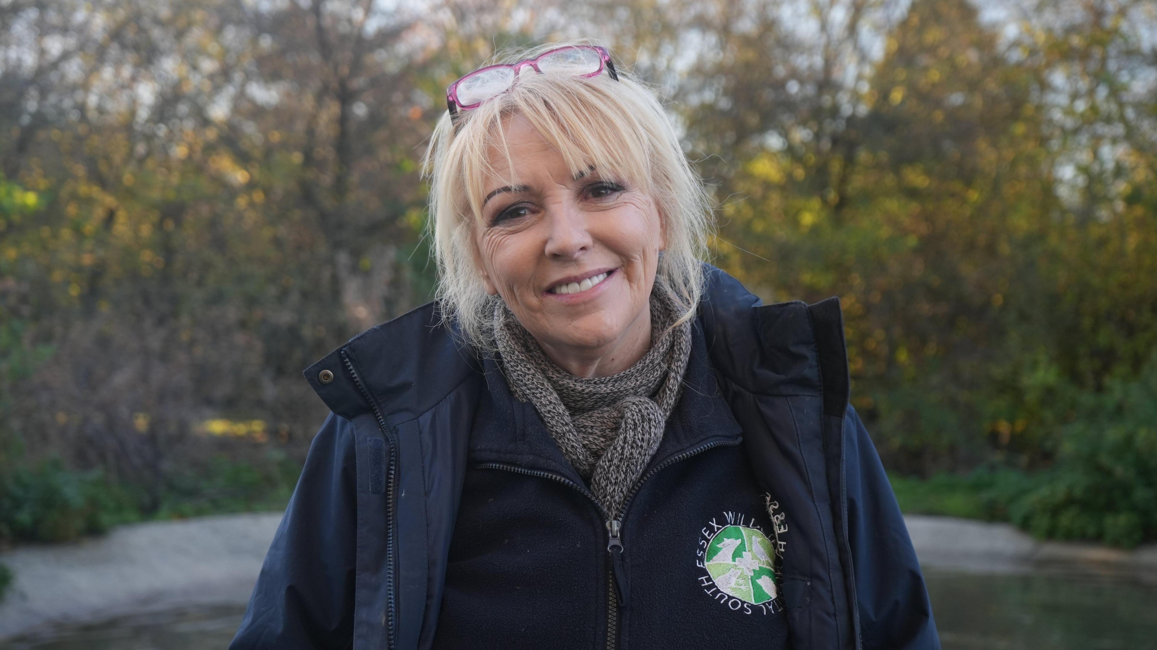 Sue Schwar is wearing a grey scarf, navy fleece and jacket with a green badge that says 'South Essex Wildlife Hospital'. Her hair is blonde and straight and she has a fringe. On top of her head sits a pair of pink glasses. She is standing in front of an outdoor pool surrounded by trees in the sunshine.