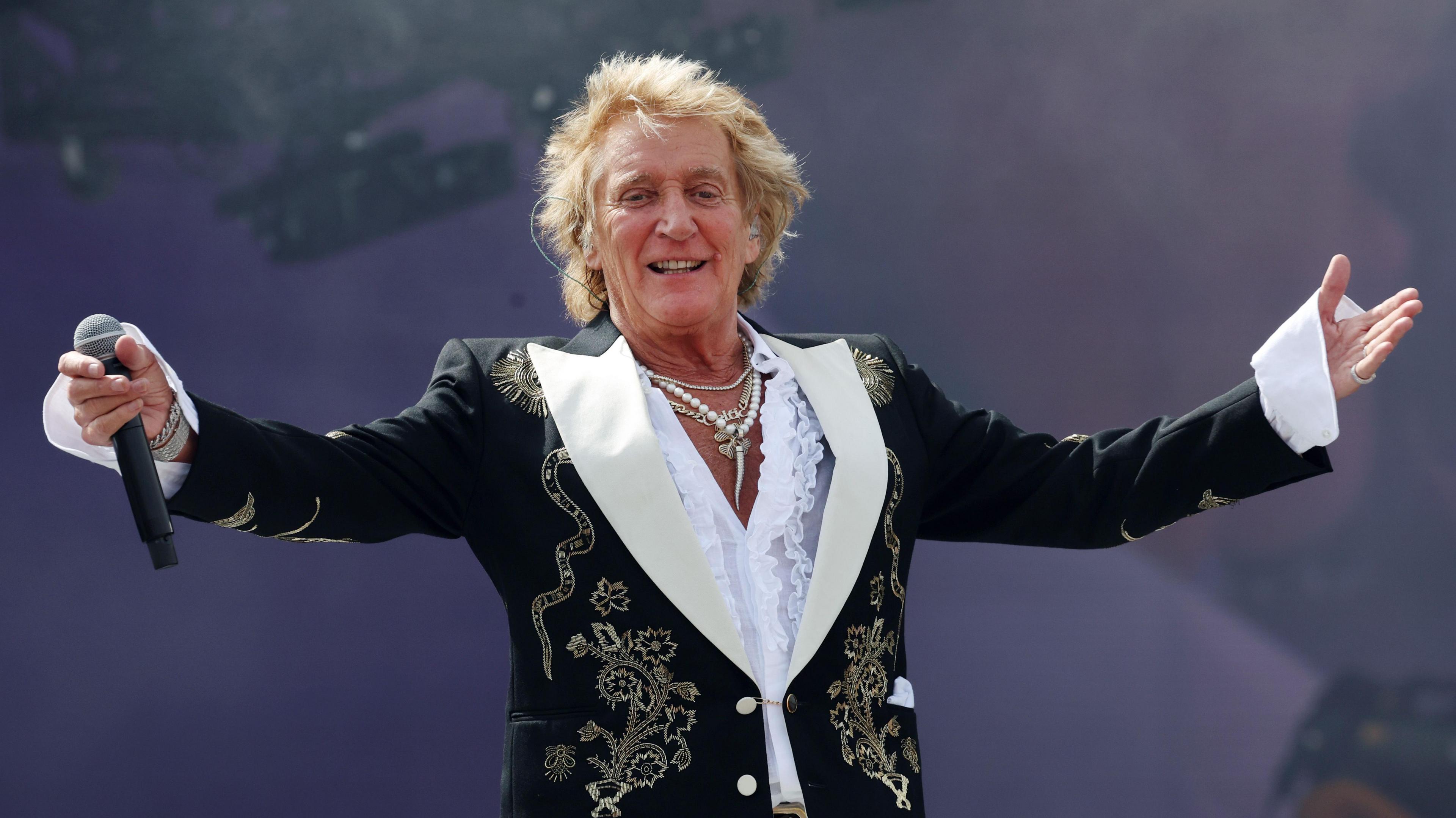 Rod Stewart is standing on stage wearing a black jacket with stitched flowers. He is spreading his arms towards the crowd and has a microphone in hand. His shirt has a low neckline and he is wearing about three necklaces.