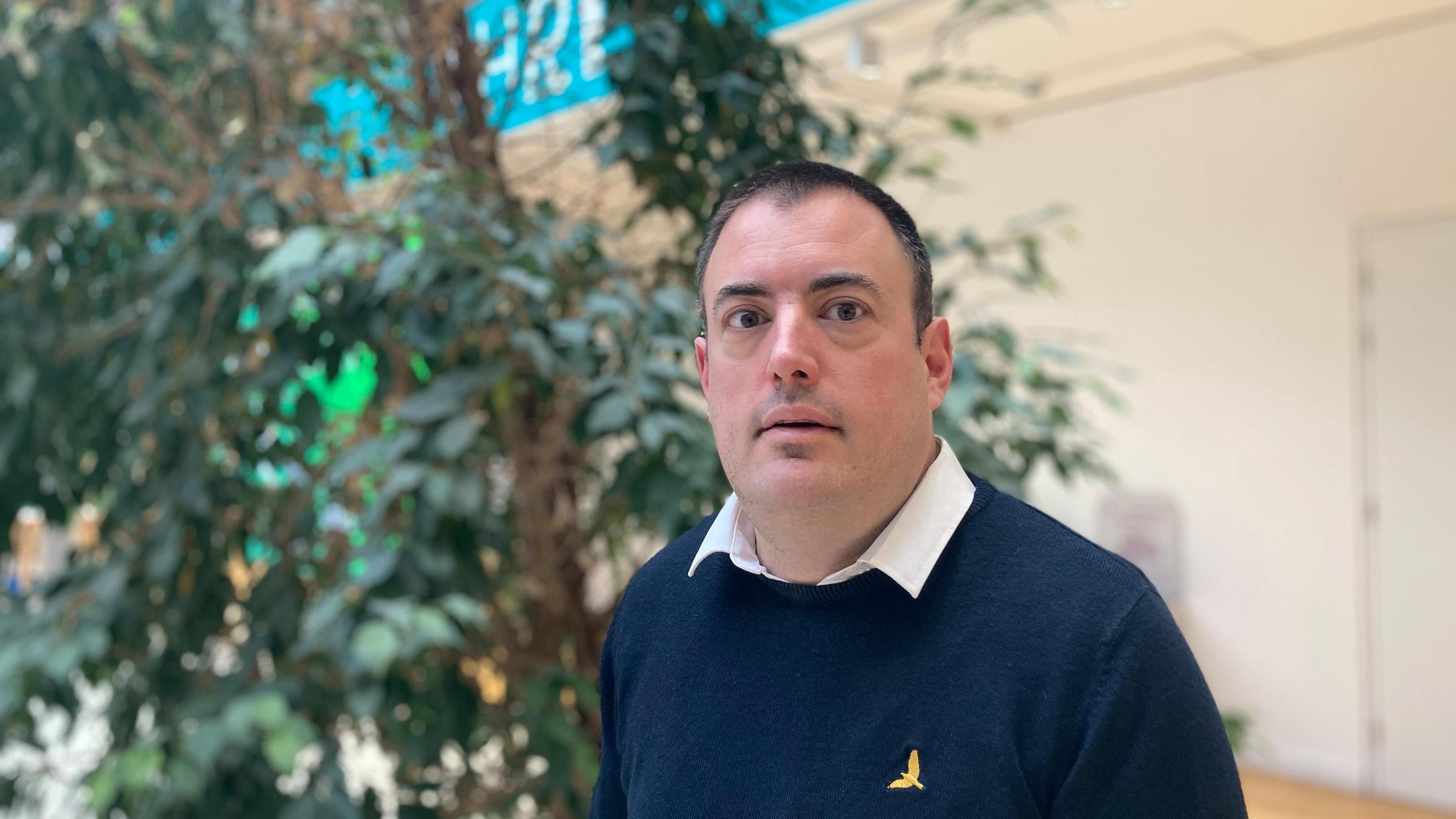 Man wearing a navy jumper and white shirt in the atrium of an office building with a plant in the background 