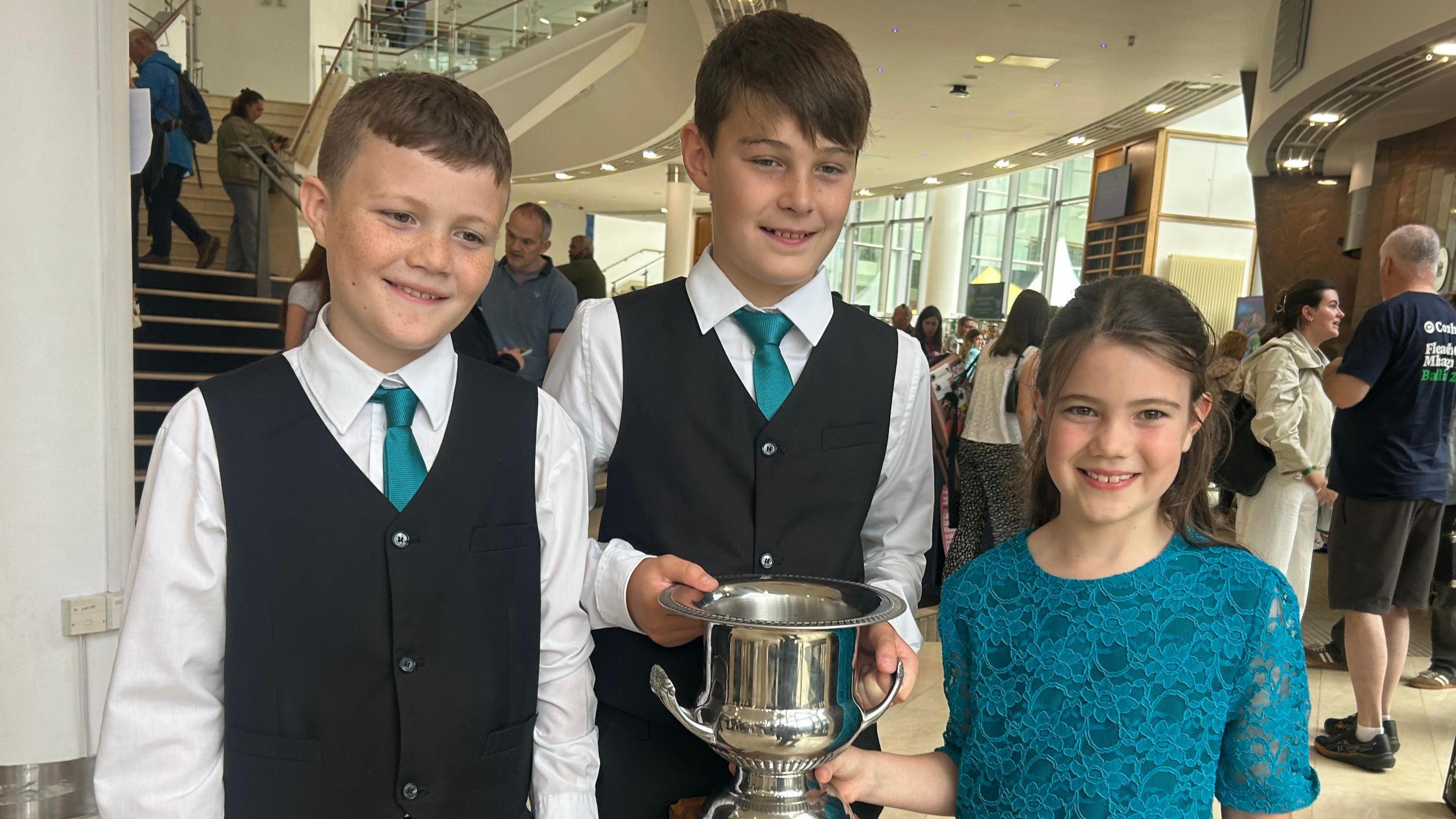 Two boys and a girl standing beside each other. The boys are wearing white shirts, blue ties and black waistcoats. The girl is wearing a blue top. The boy in the middle is holding a silver trophy. 