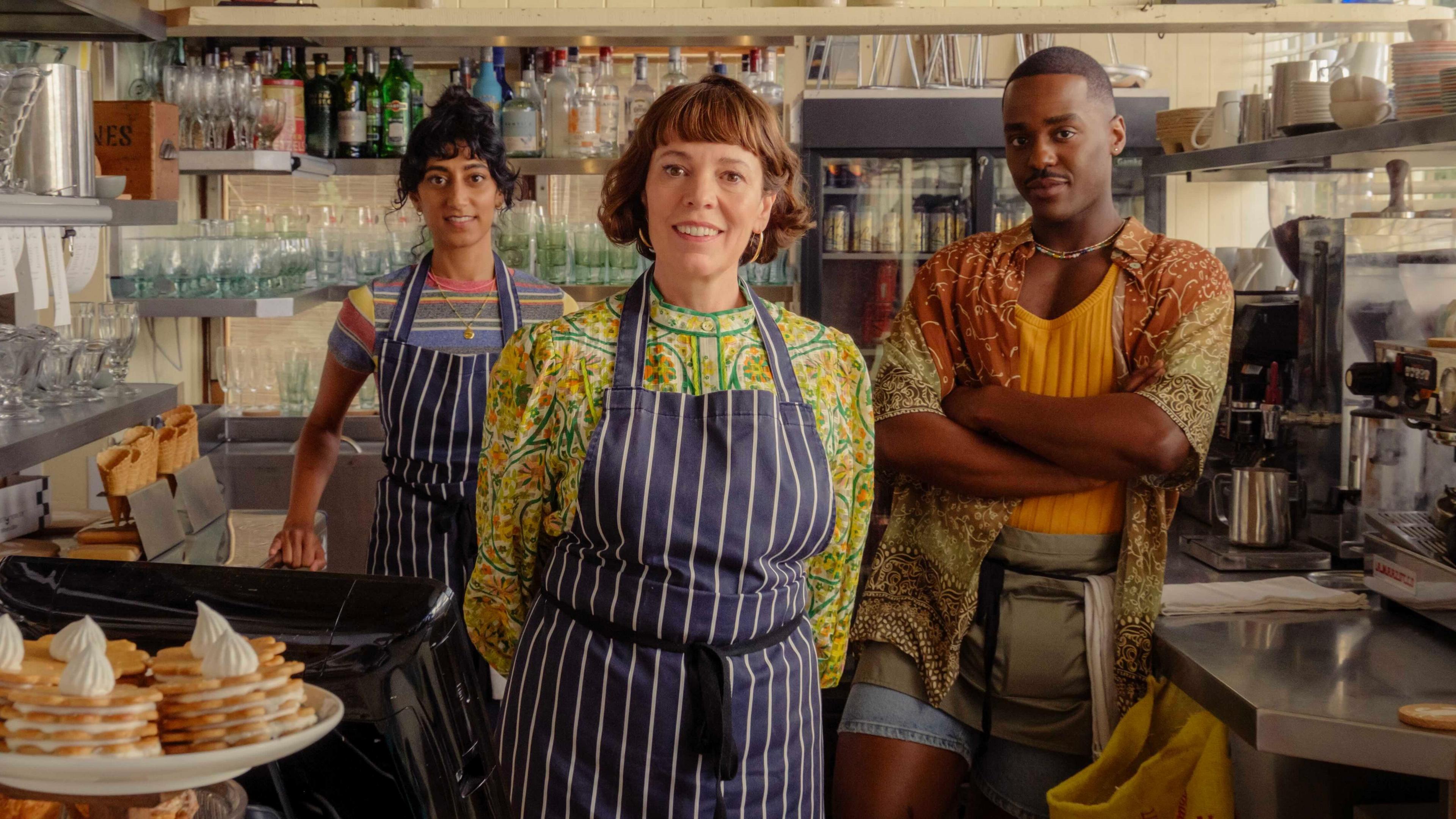 The image shows three individuals standing behind the counter of a café, all wearing aprons. The counter is filled with items like plates, glasses, a coffee machine, and a display of pastries. Shelves behind them are stocked with bottles and glassware, and a menu board above lists drinks and food options.
