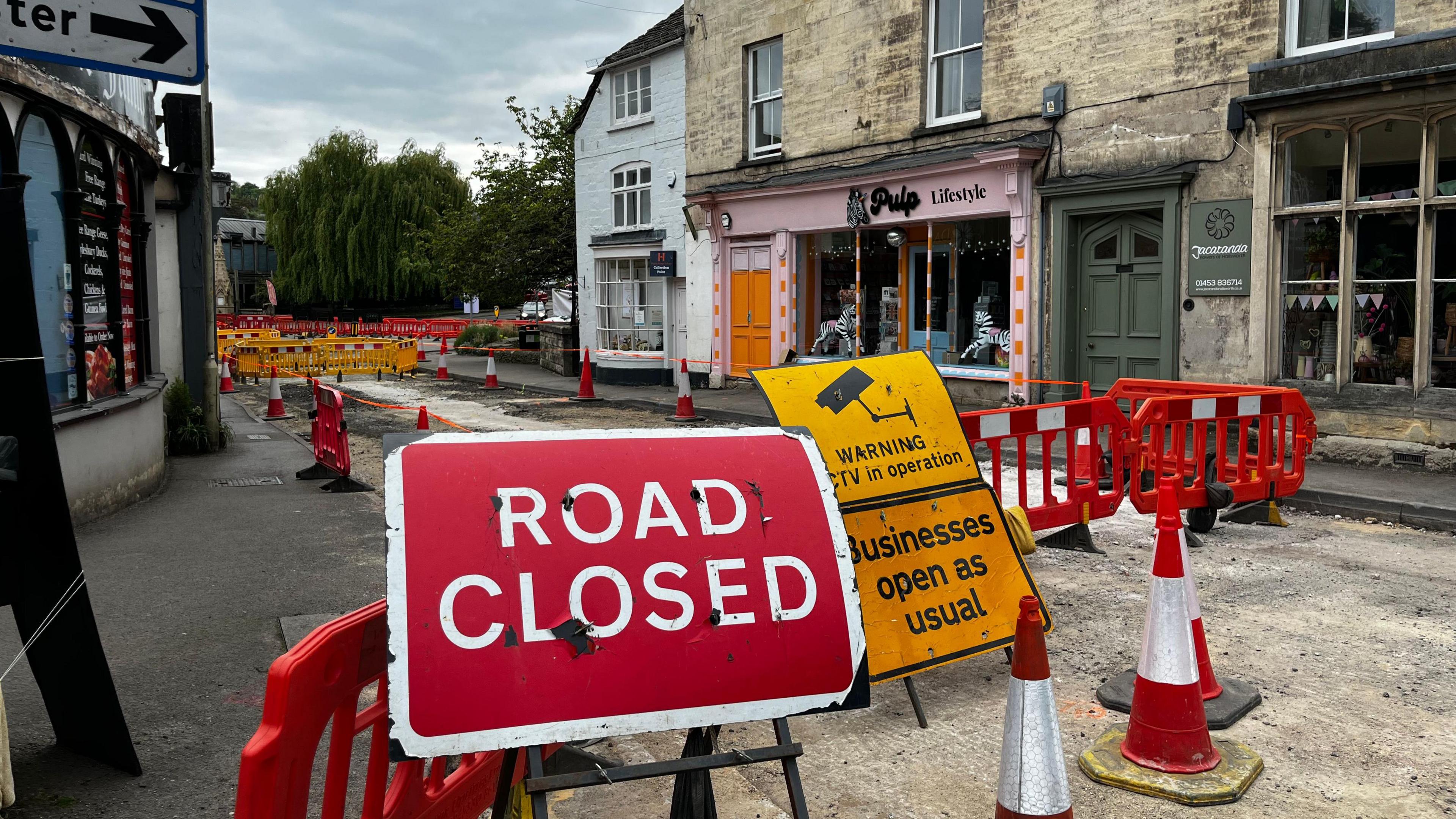 A road closure signed in front of fencing and cones while work is carried out to the road. Behind are local shops.