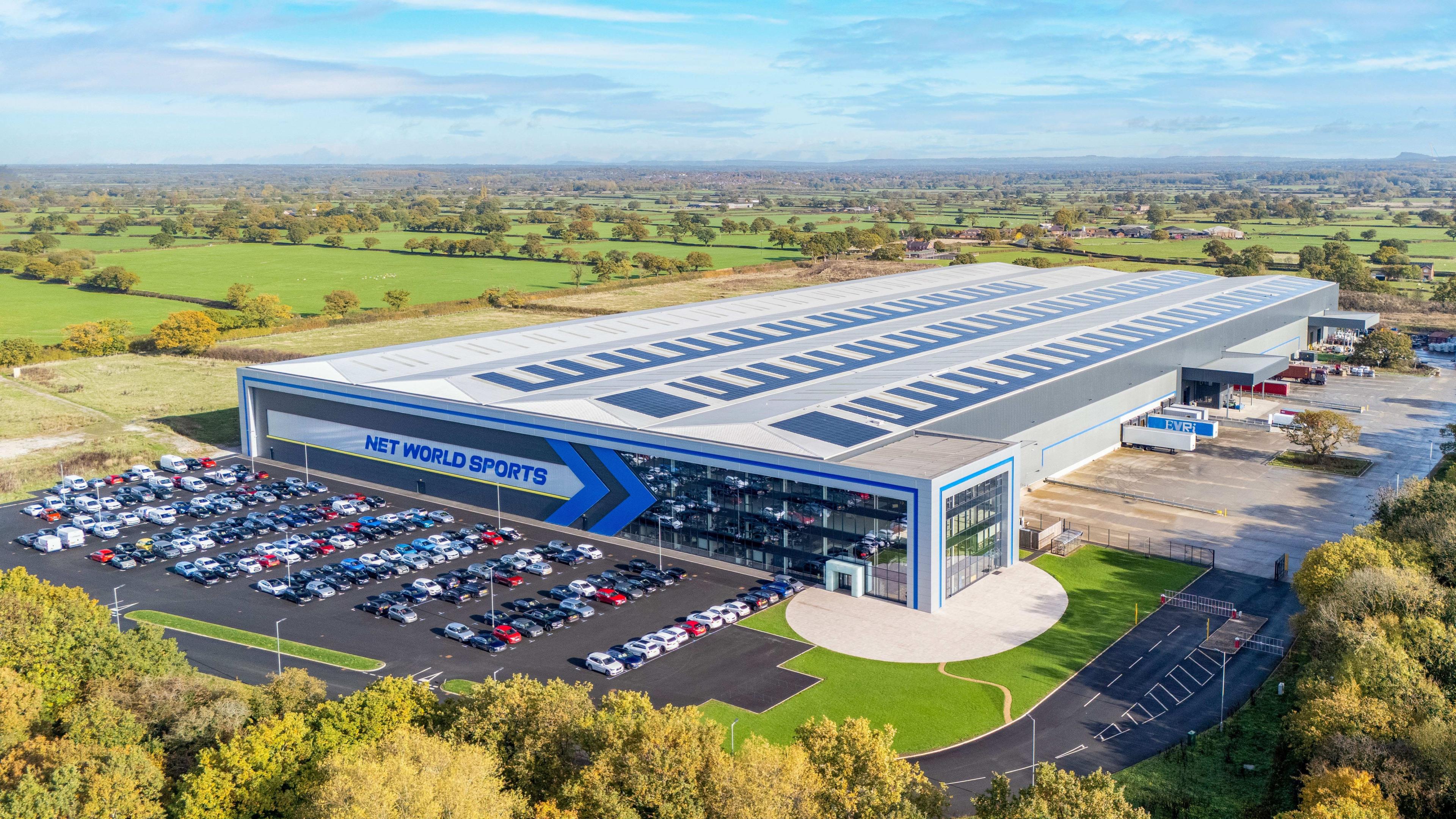 Adeilad diwydiannol mawr, modern gyda ffenestri mawr ar y blaen a’r geiriau “NET WORLD SPORTS”  yn amlwg ar ochr y strwythur mewn llythrennau glas. Mae’r adeilad yn cynnwys warws helaeth gyda rhesi o baneli solar ar y to. Mae maes parcio mawr yn llawn ceir, ac mae ffyrdd tarmac yn arwain at y prif fynedfa. Mae’r safle wedi’i amgylchynu gan goed a thir amaethyddol gwyrdd, gyda golygfeydd eang o gaeau yn ymestyn i’r gorwel o dan awyr las gyda chymylau.