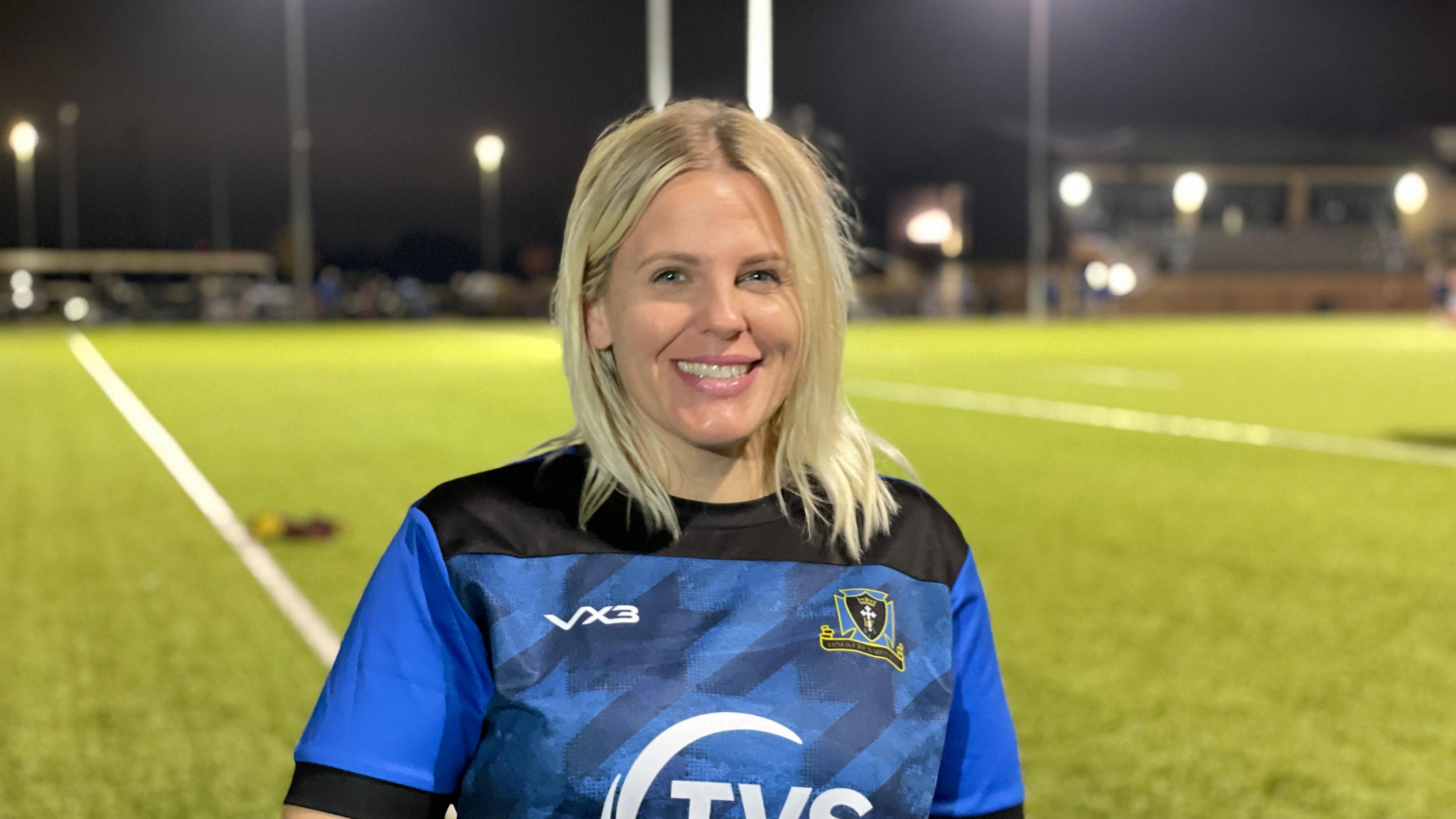 Rebecca Bird smiles at the camera. She has blonde hair down to her shoulders and is wearing a blue and black rugby shirt with the Dings Crusaders RFC logo on the right side. Behind her is a blurred image of a rugby pitch.