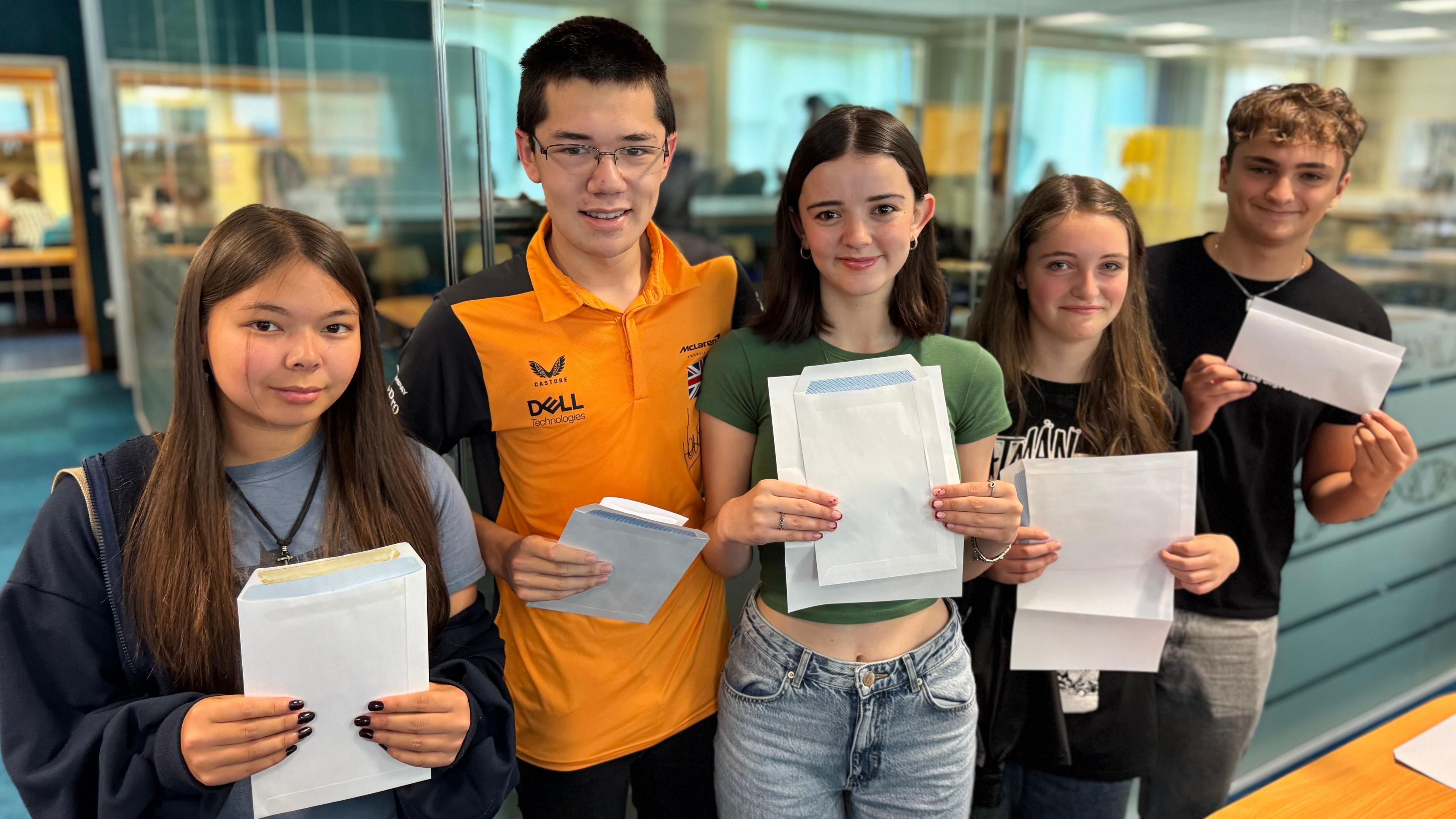 A group of teenage boys and girls stand together and smile in front of a glass classroom. They wear casual clothes and hold up envelopes.