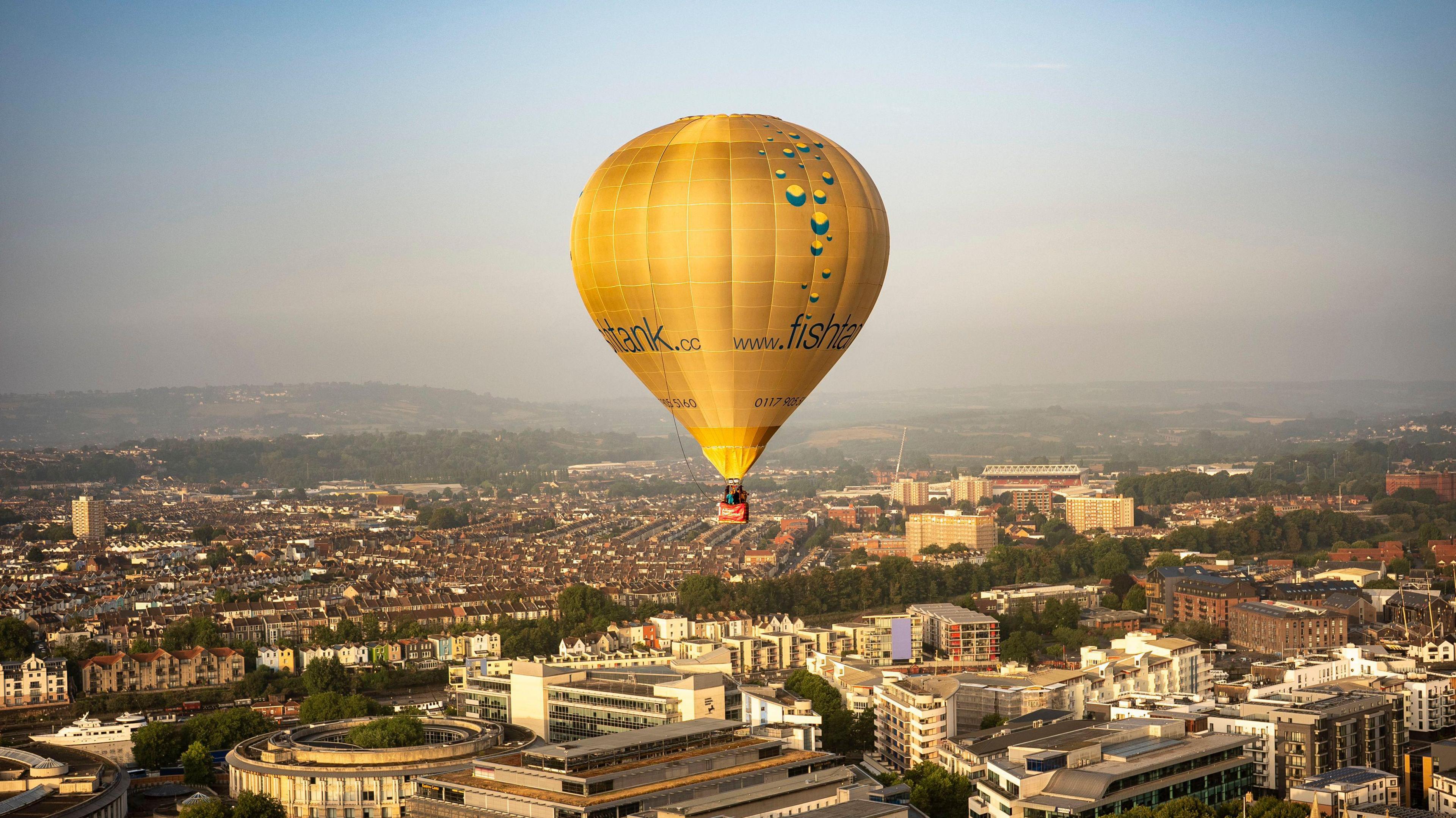 A gold hot air balloon flying over Bristol. The city landscape can be seen below. 