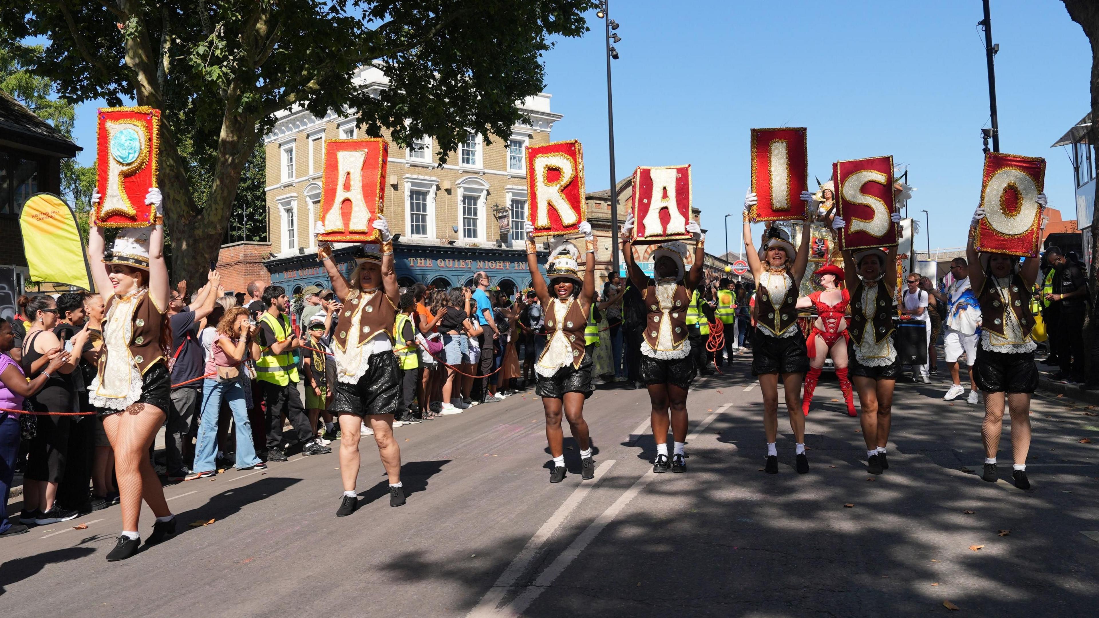 Performers hold up the letters P, A, R, A, I, S and O which are written on red backgrounds as they parade along a street with crowds of people along the road