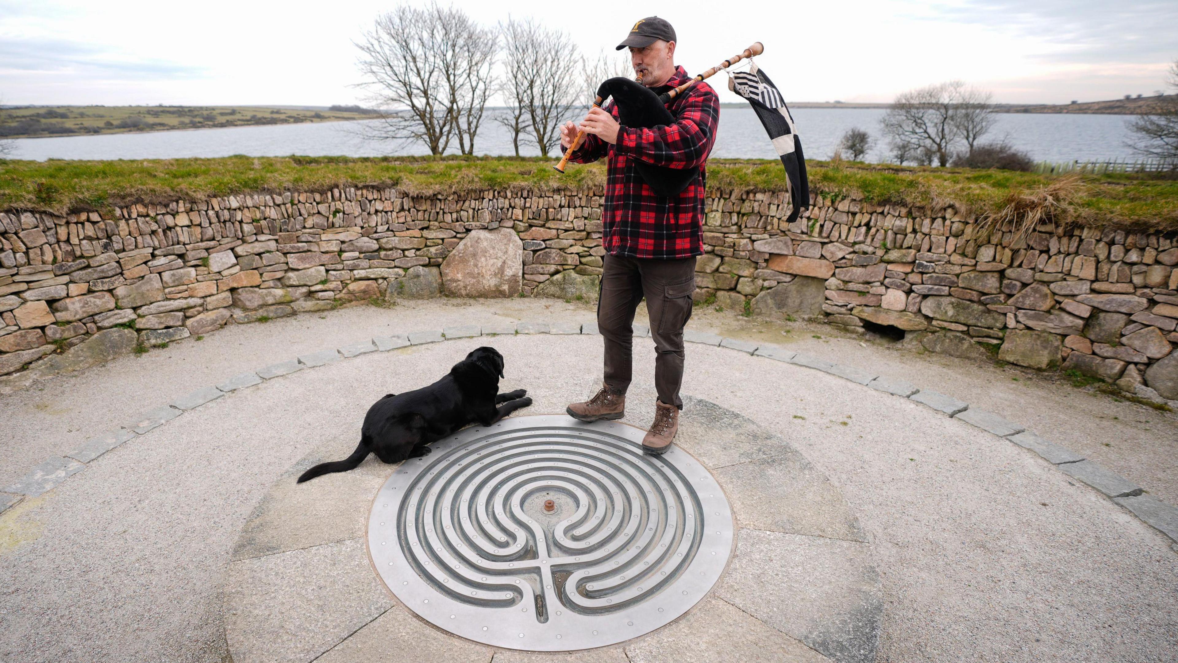 A picture of a man and a dog stood in the middle of the labrynth playing an instrument with the Cornish flag waving behind him. His dog is sat down next to him. They are surrounded by a solid wall with trees and a bed of water. 