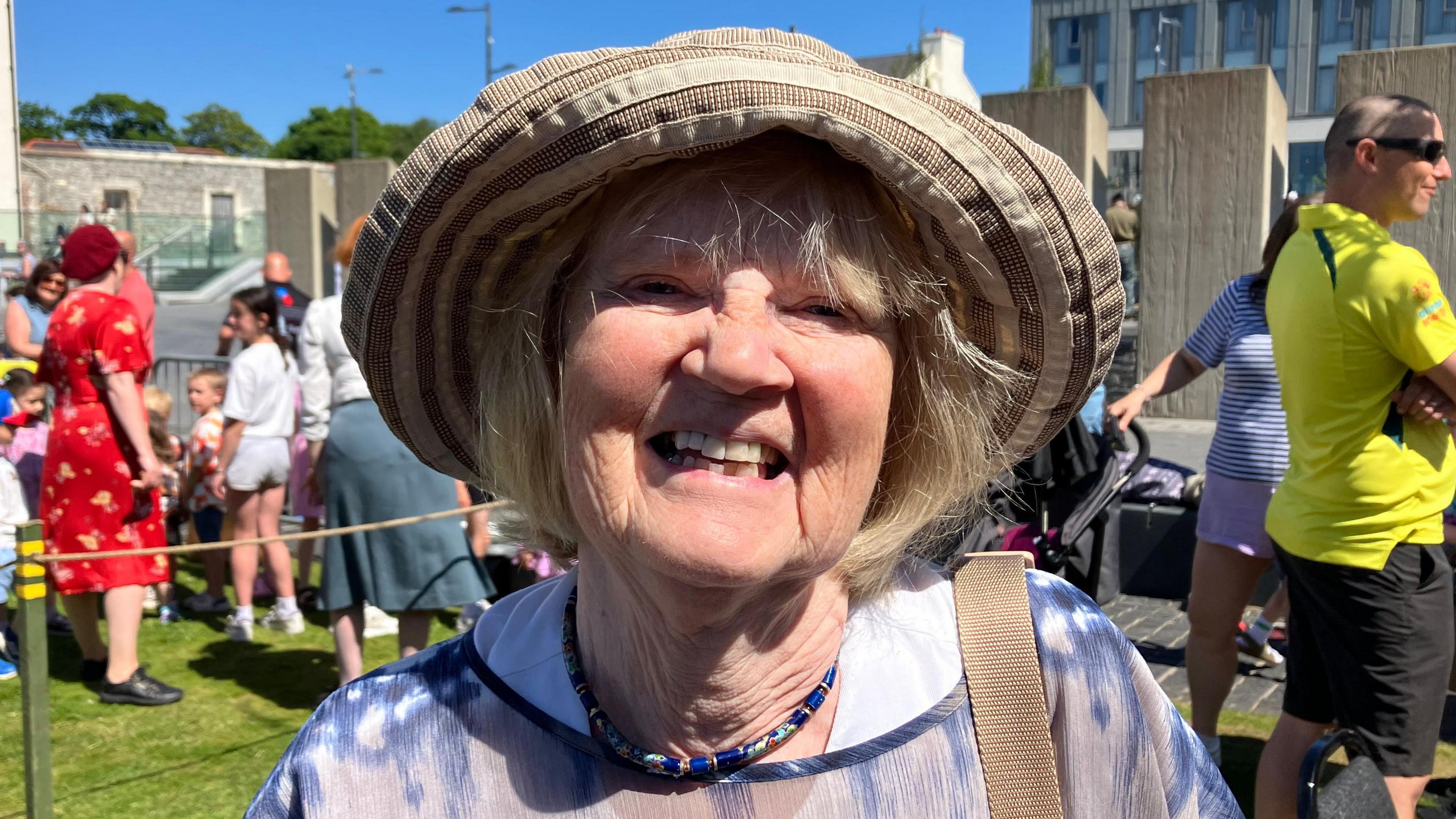 Rhoda Baxter is wearing a brown and weight hat, she has a strap over her left shoulder. She is wearing a beaded necklace and a blue and white shirt. Behind her is a crowd of people and a number of large stone pieces.