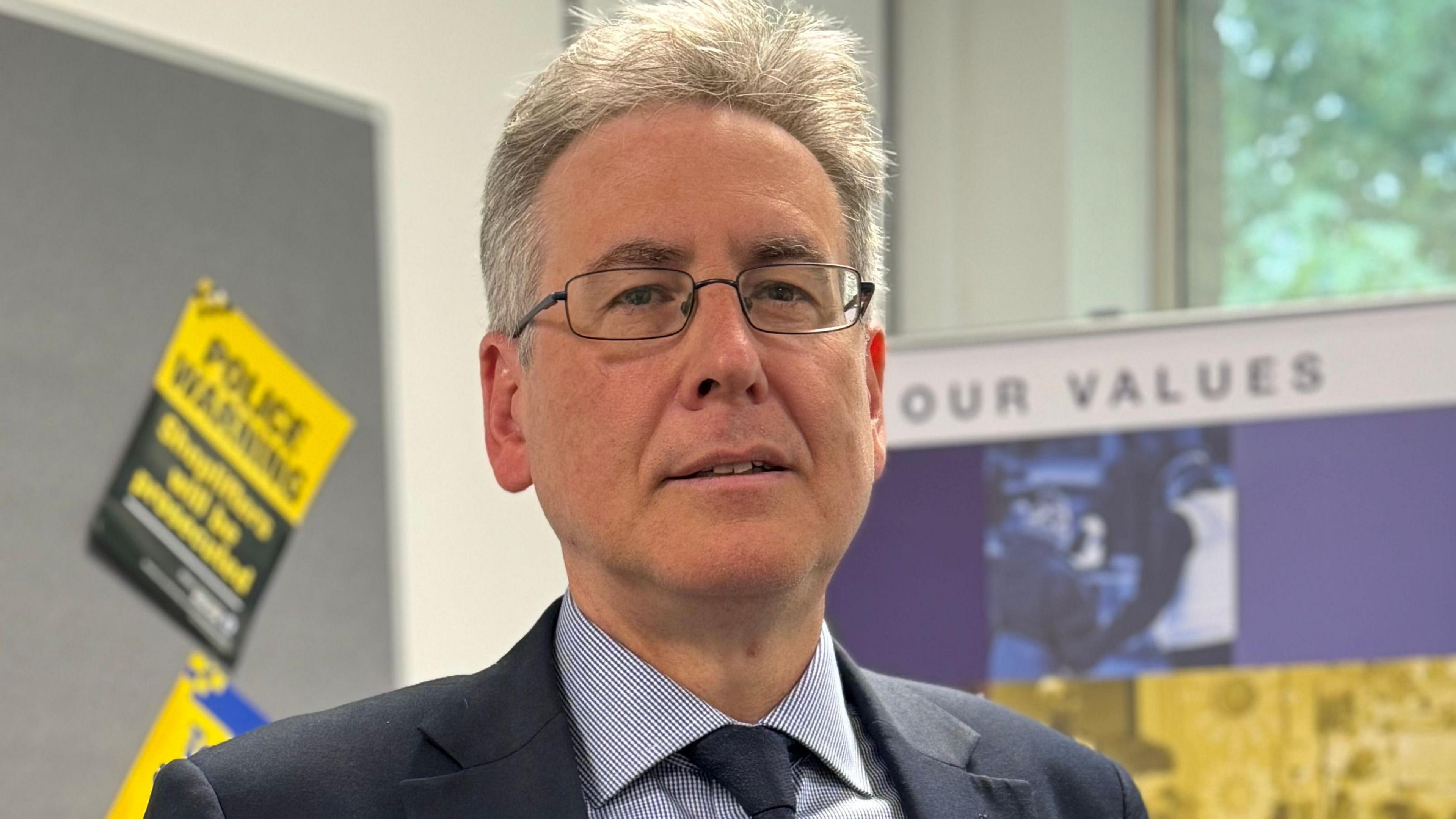 Një burrë me flokë të bardhë dhe syze me kornizë teli në një dhomë me fletëpalosje policie të ngjitura në tabela njoftimesh. Ai ka veshur një kostum blu të errët dhe një këmishë blu me kuadrata.