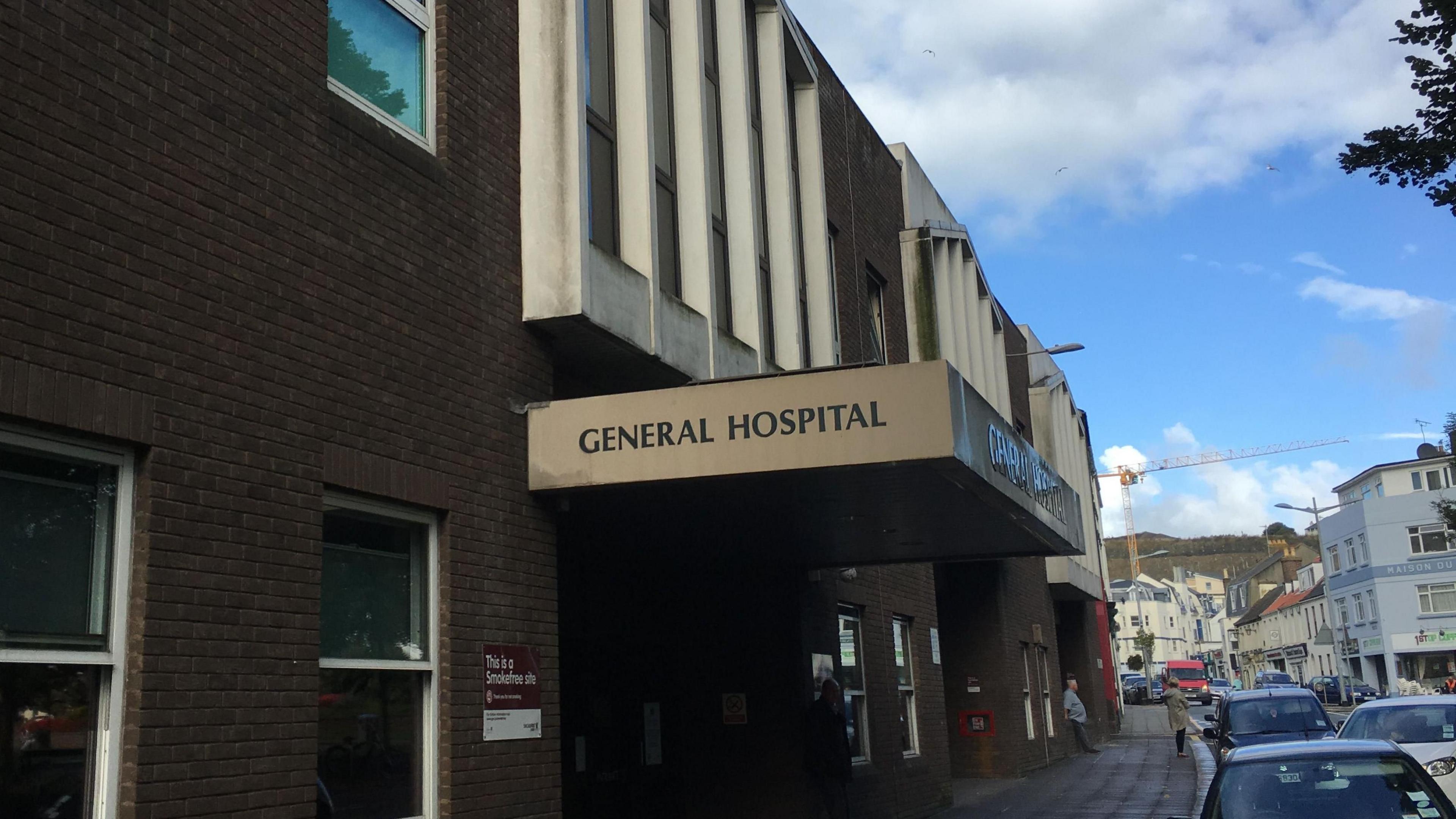 A picture of the outside of Jersey General Hospital with a pavement outside, parked cars and a road