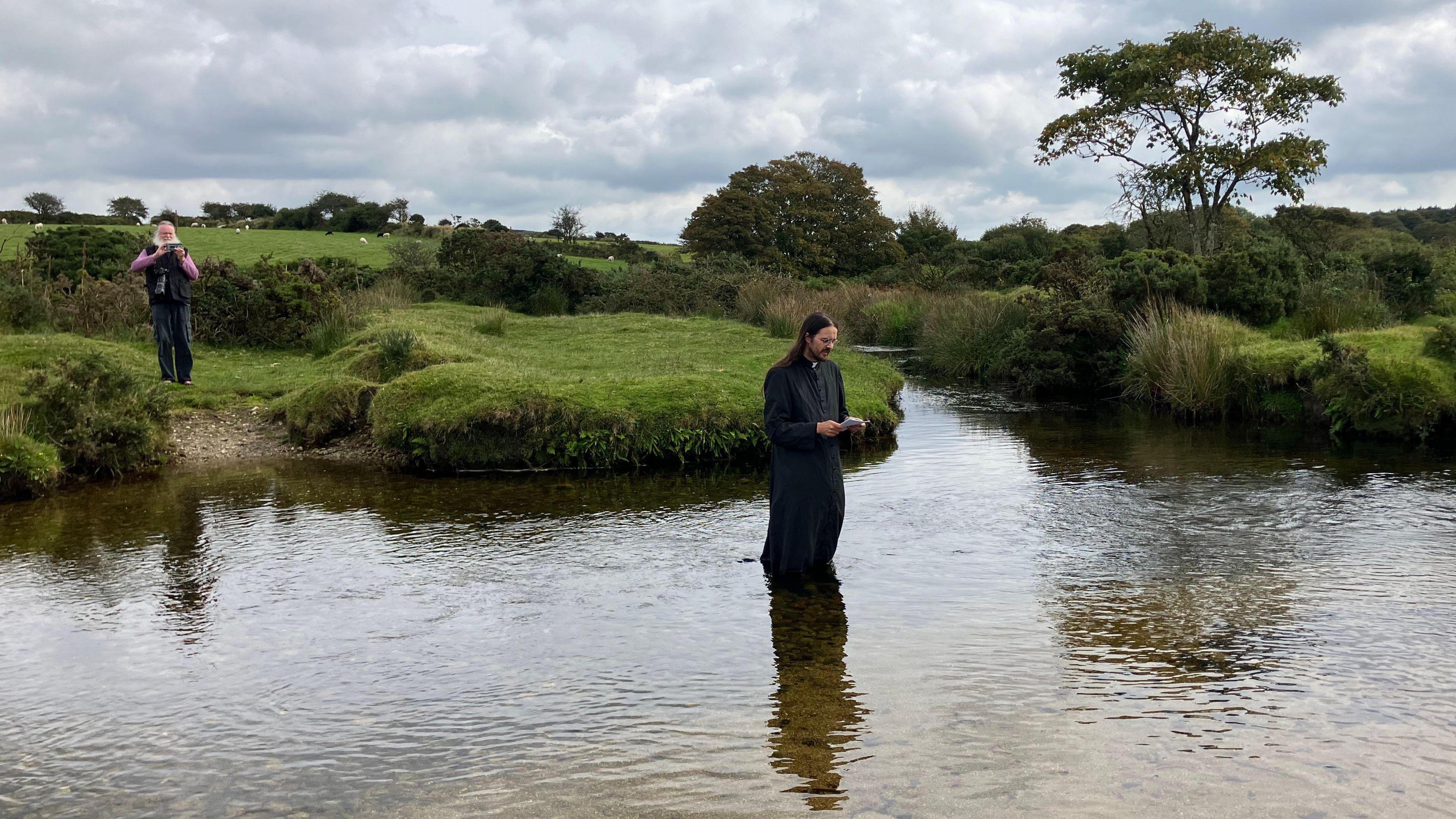 Reverend Robin Thwaites in the river on Bodmin Moor, wearing robes and holding a book, while a man with a white beard overlooks from the far, grassy bank