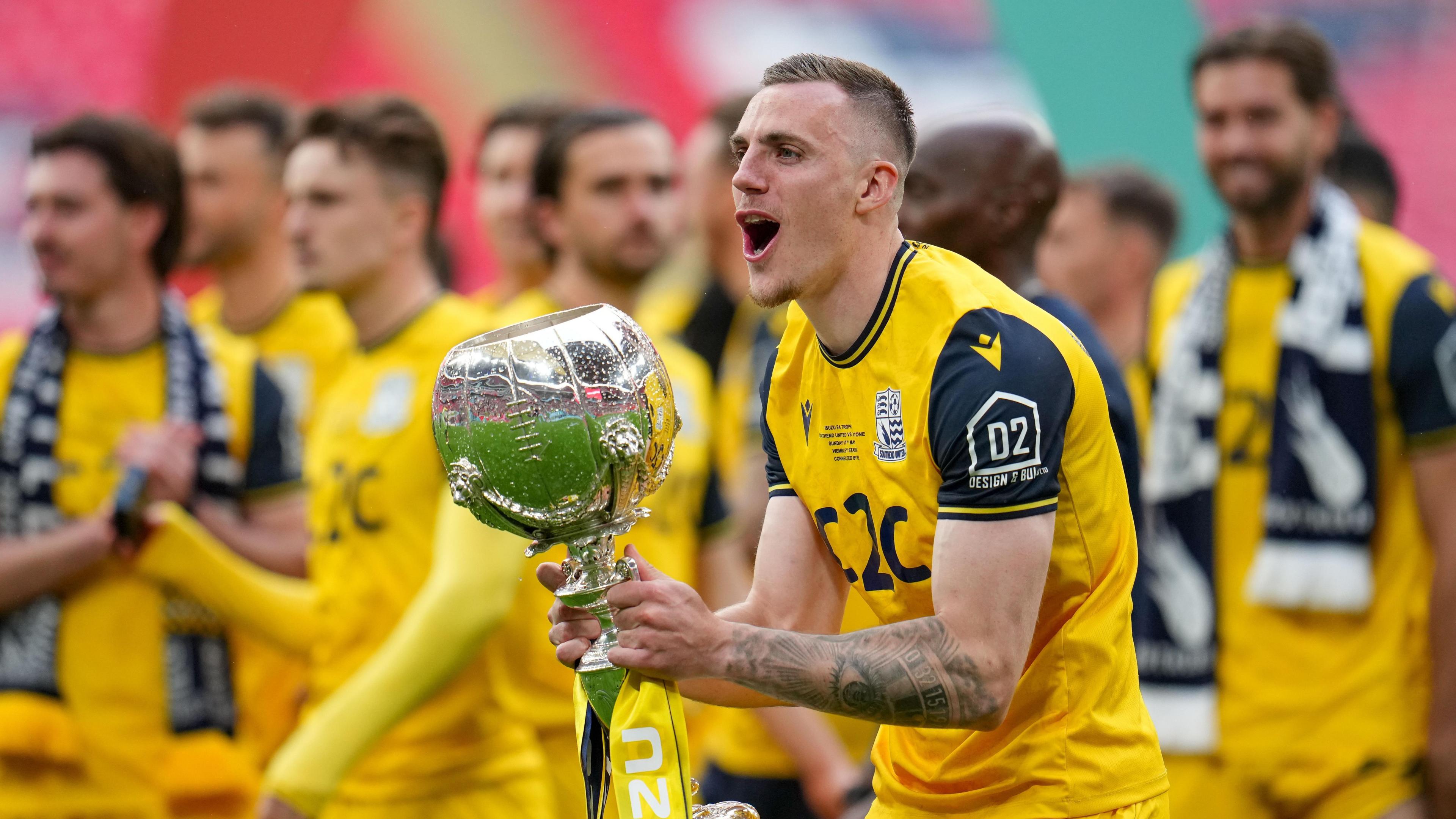 Southend United defender Ben Goodliffe clutching the FA Trophy after defeating Wealdstone in May with his team-mates slightly blurred in the background