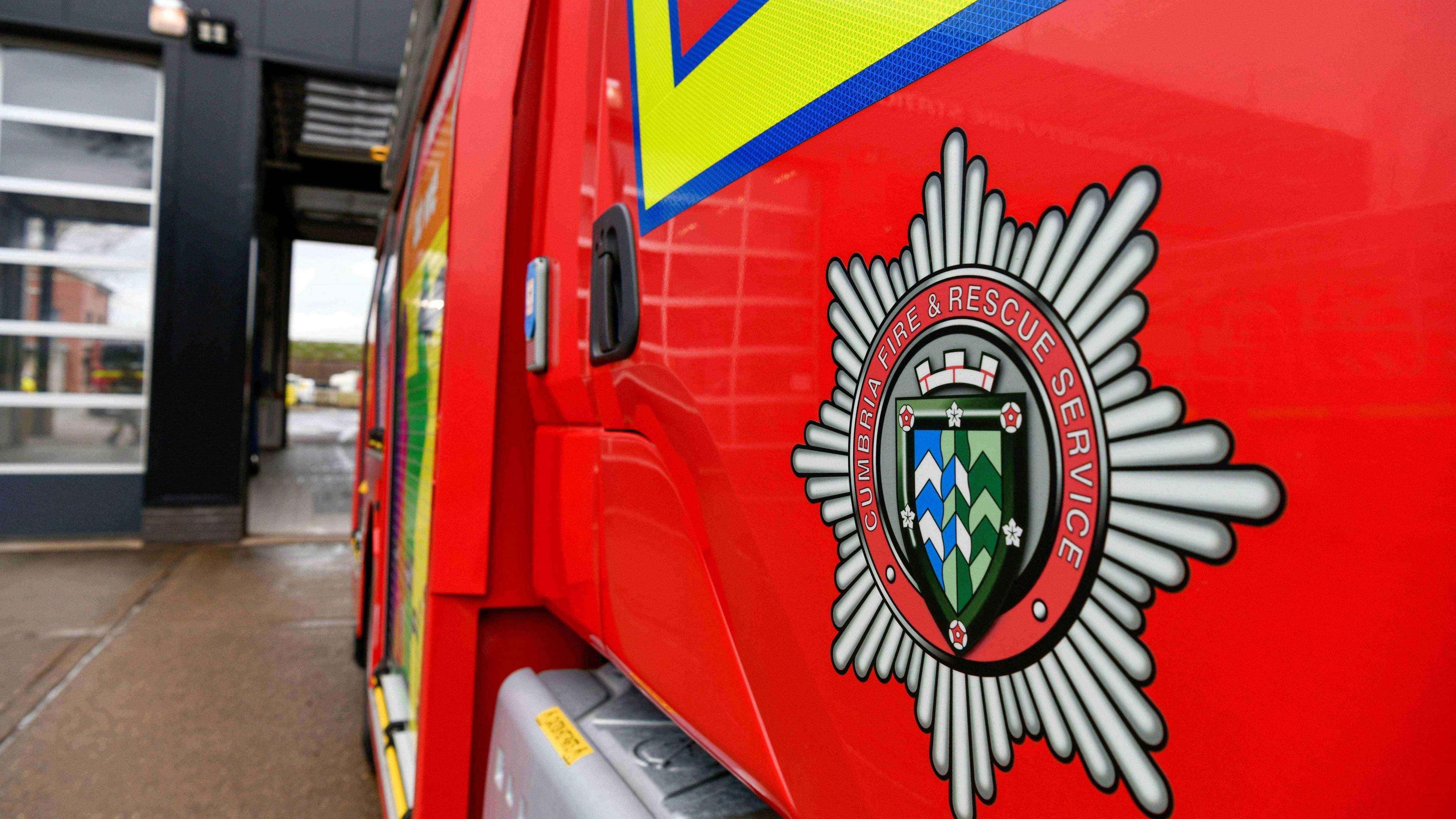 A stock image of a fire engine. The red vehicle is pictured from the side, focusing on the badge with the name Cumbria Fire and Rescue Service, with an emblem in the shape of a shield, with a crown on the top.