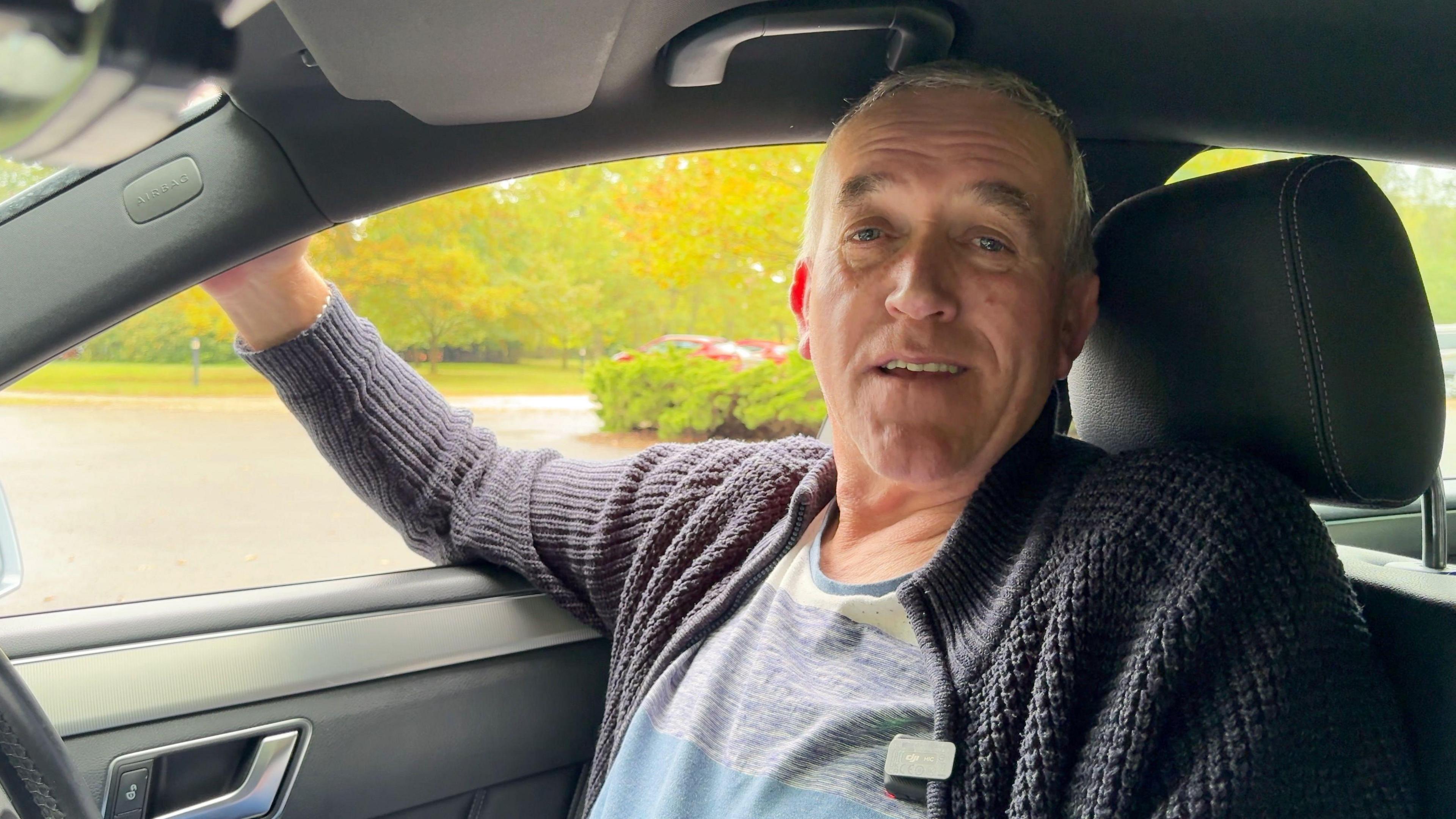 Taxi driver Steve pictured in his taxi, he is wearing a striped t-shirt and blue woolen cardigan. He has one arm out of the side of his taxi window. 