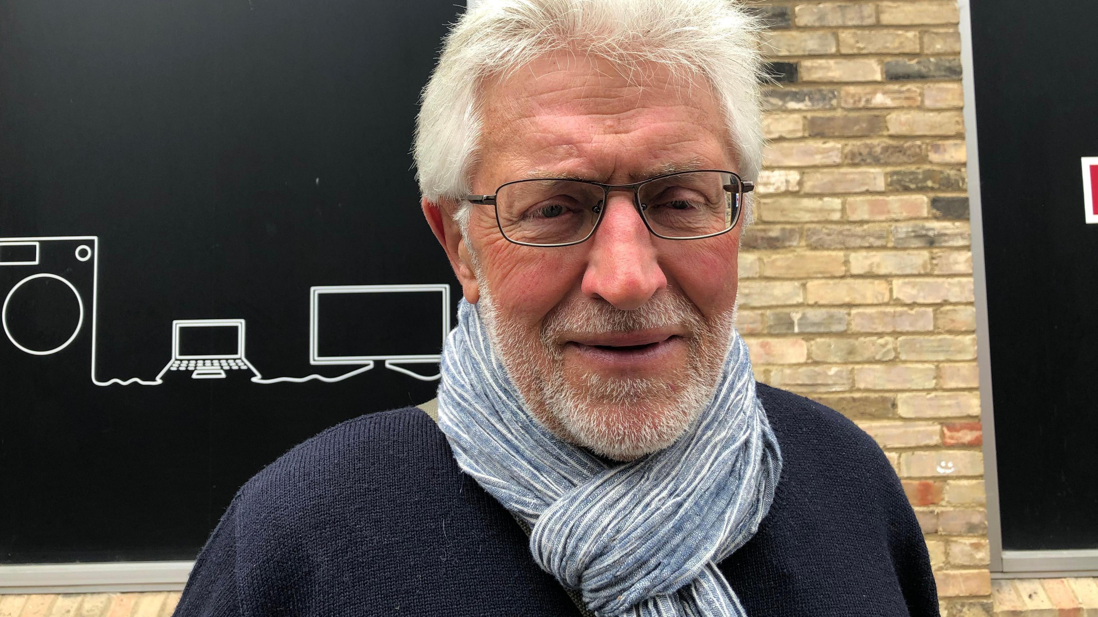 A man wearing a blue jumper and with a blue & white scarf looped around his neck wearing glasses