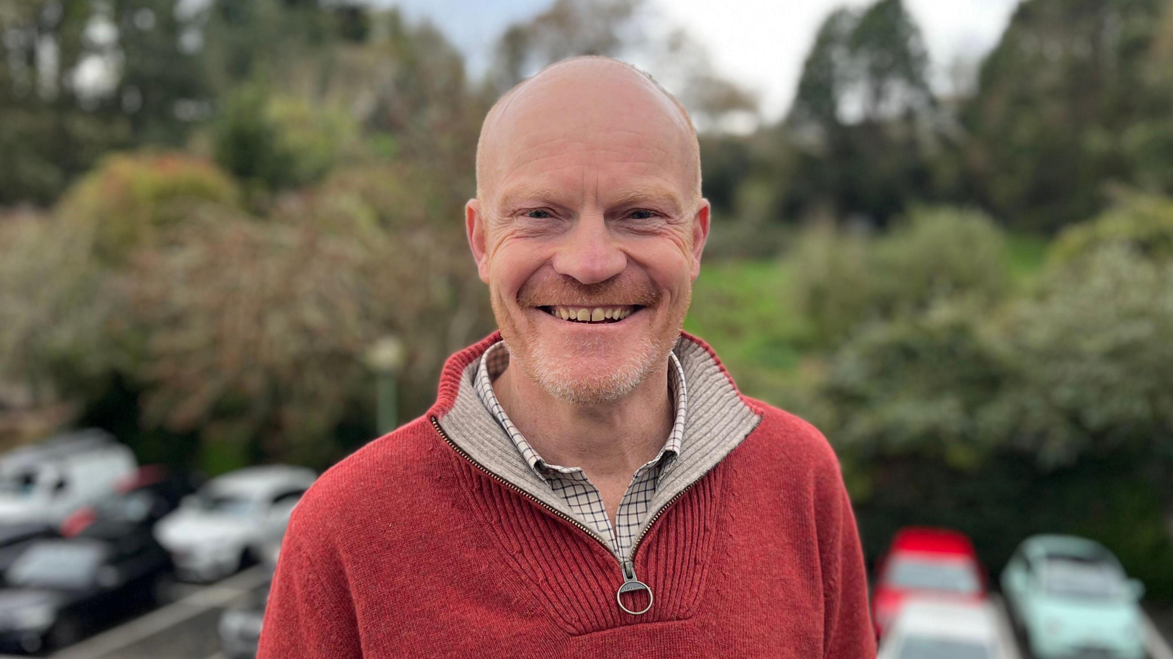 Deputy Gavin St Pier - A man with a bald head and a ginger and grey beard smiling at the camera in a car park. He is wearing a red jumper.
