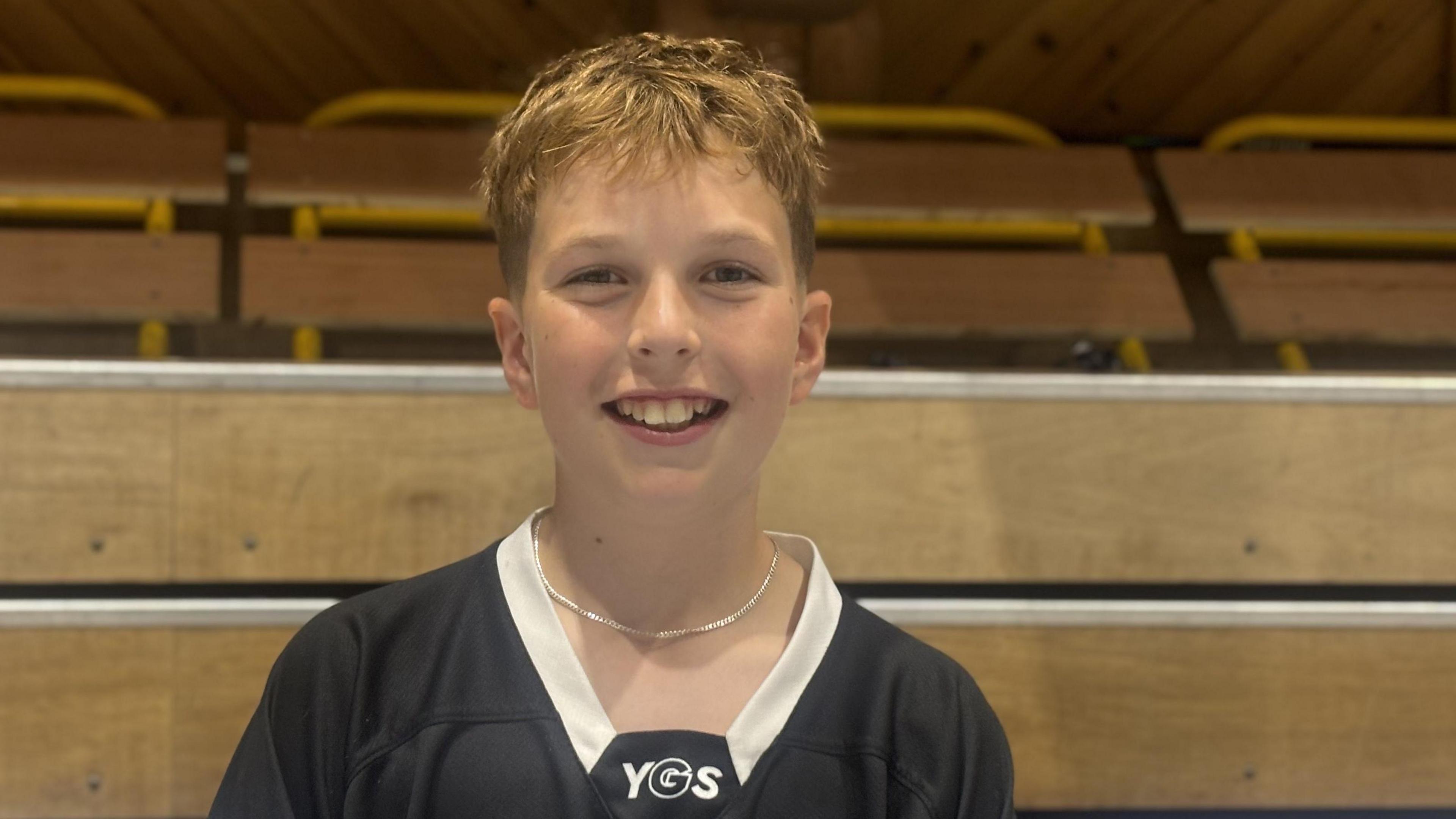 Archie is a young, fair-skinned boy with blonde hair wearing a black jersey with white on the neck. He is standing in the roller rink with a side wall behind him.