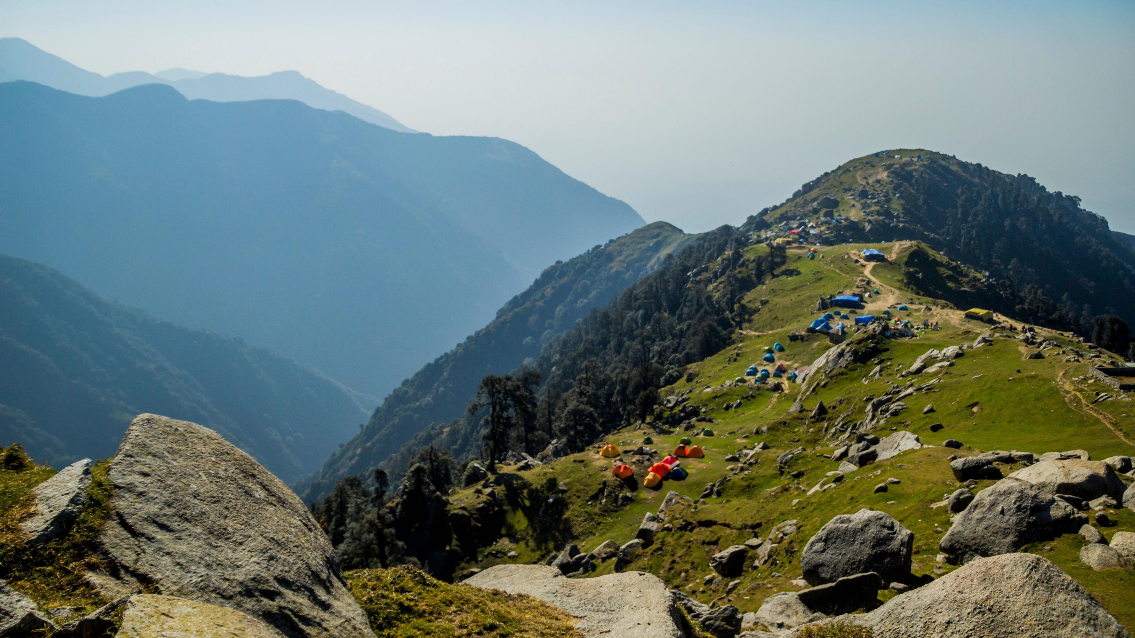 A grass-topped mountain, with some small tents dotted along, and some mountains in the background