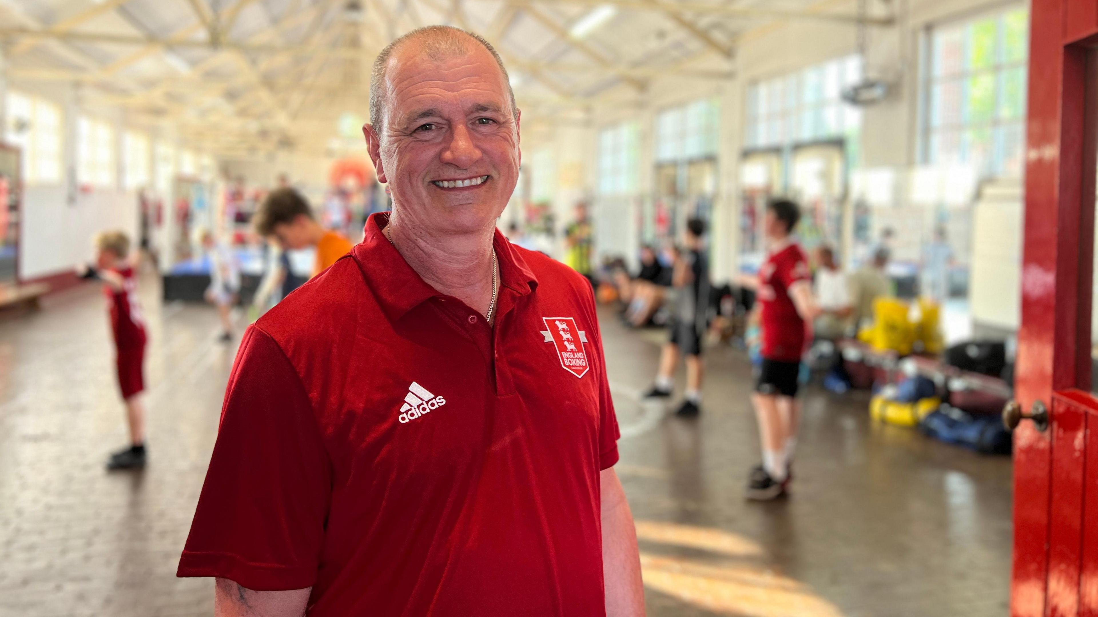 Richard is in a red polo shirt with an embroidered logo and Adidas emblem stands in the foreground of a spacious indoor area with high ceilings and exposed beams. In the background, several children in matching red sportswear participate in various activities. The room is brightly lit by natural light streaming through large windows on both sides