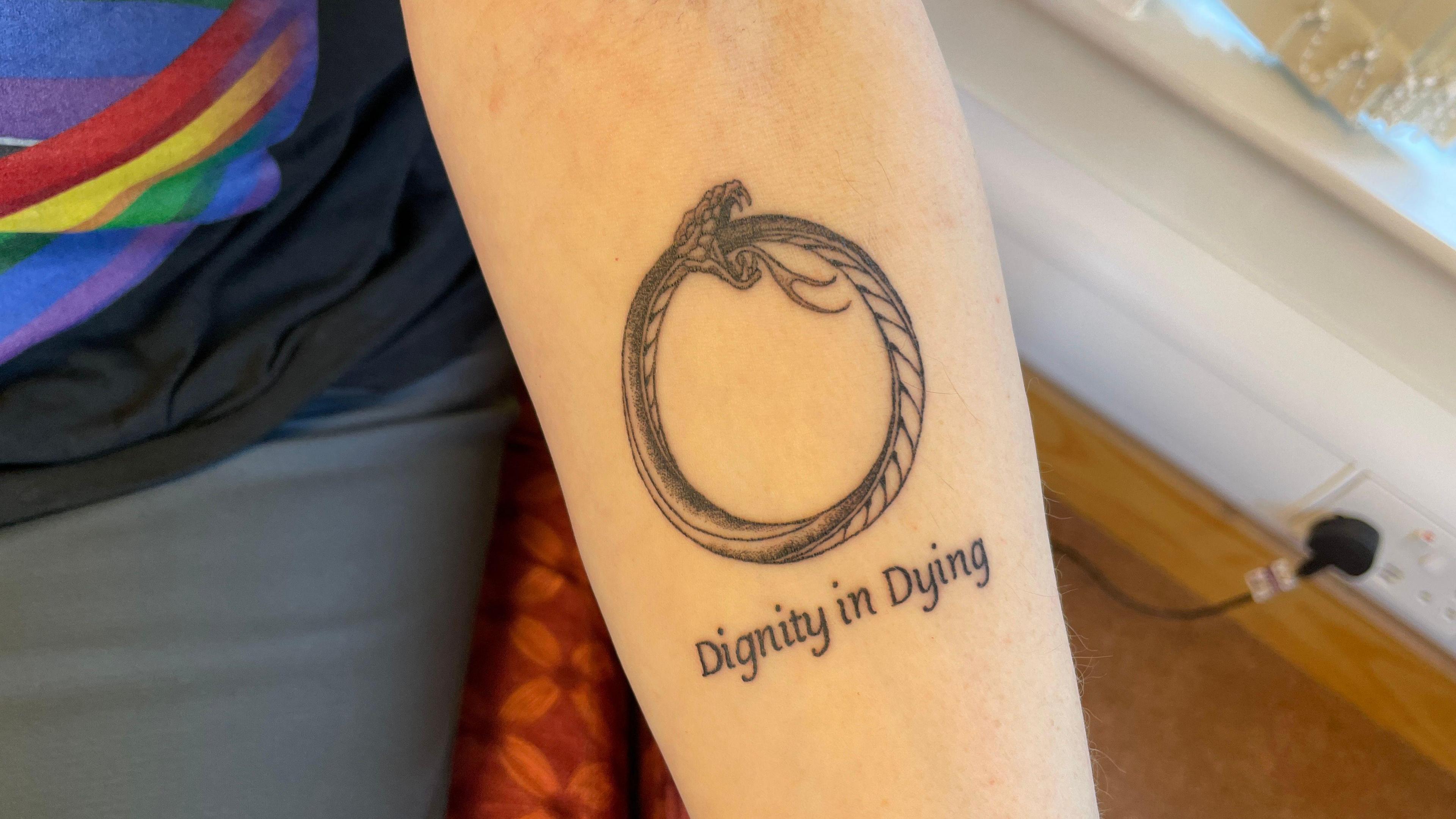 A woman's forearm with a tattoo of a snake. Underneath the image are the words "dignity in dying". The woman is wearing a black T-shirt with a rainbow printed on it and blue trousers.