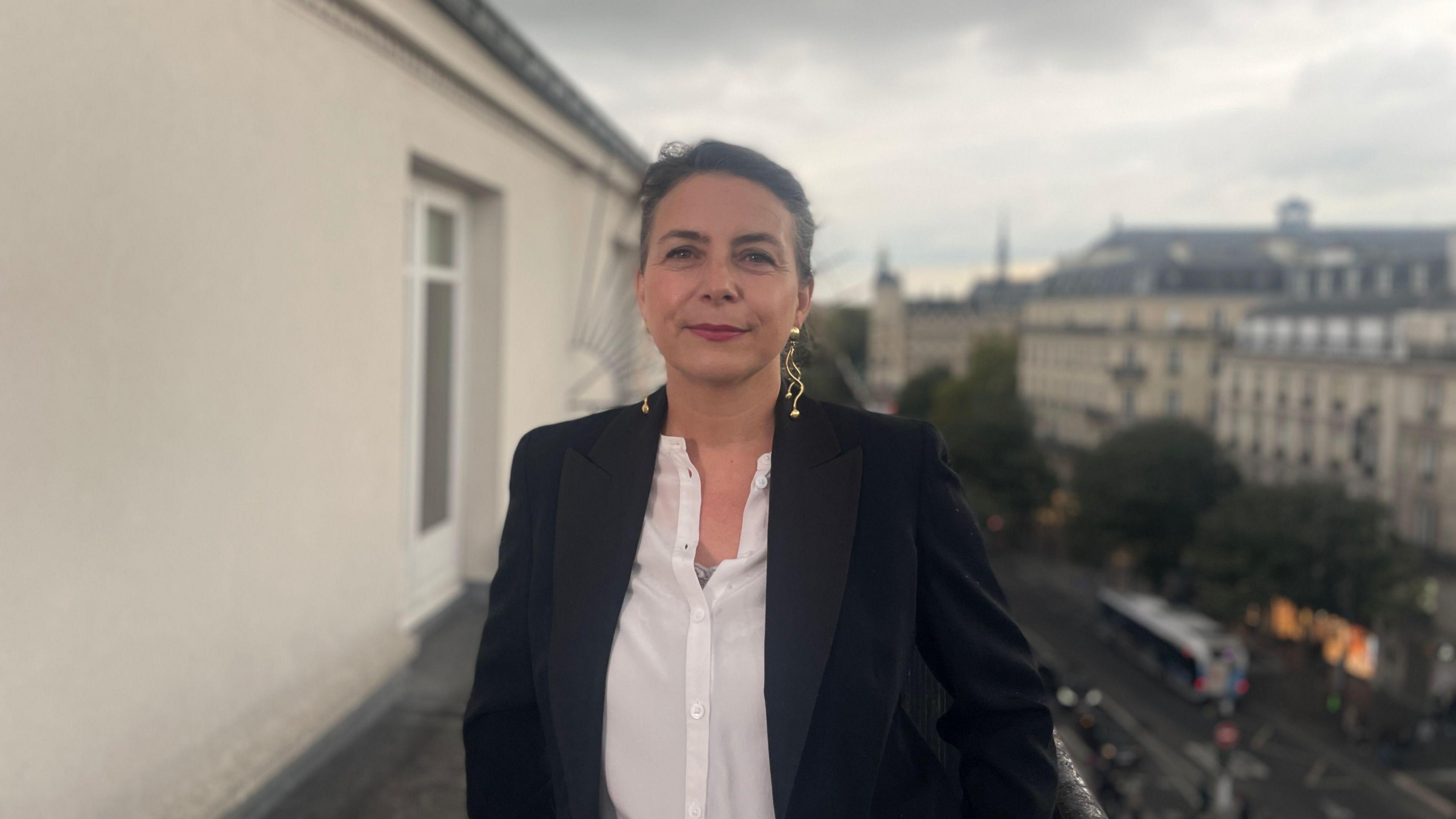 Elodie Tuaillon-Hibon is photographed on a balcony in Paris. She is looking straight into the camera and wearing a white shirt and black jacket with large gold earrings. 