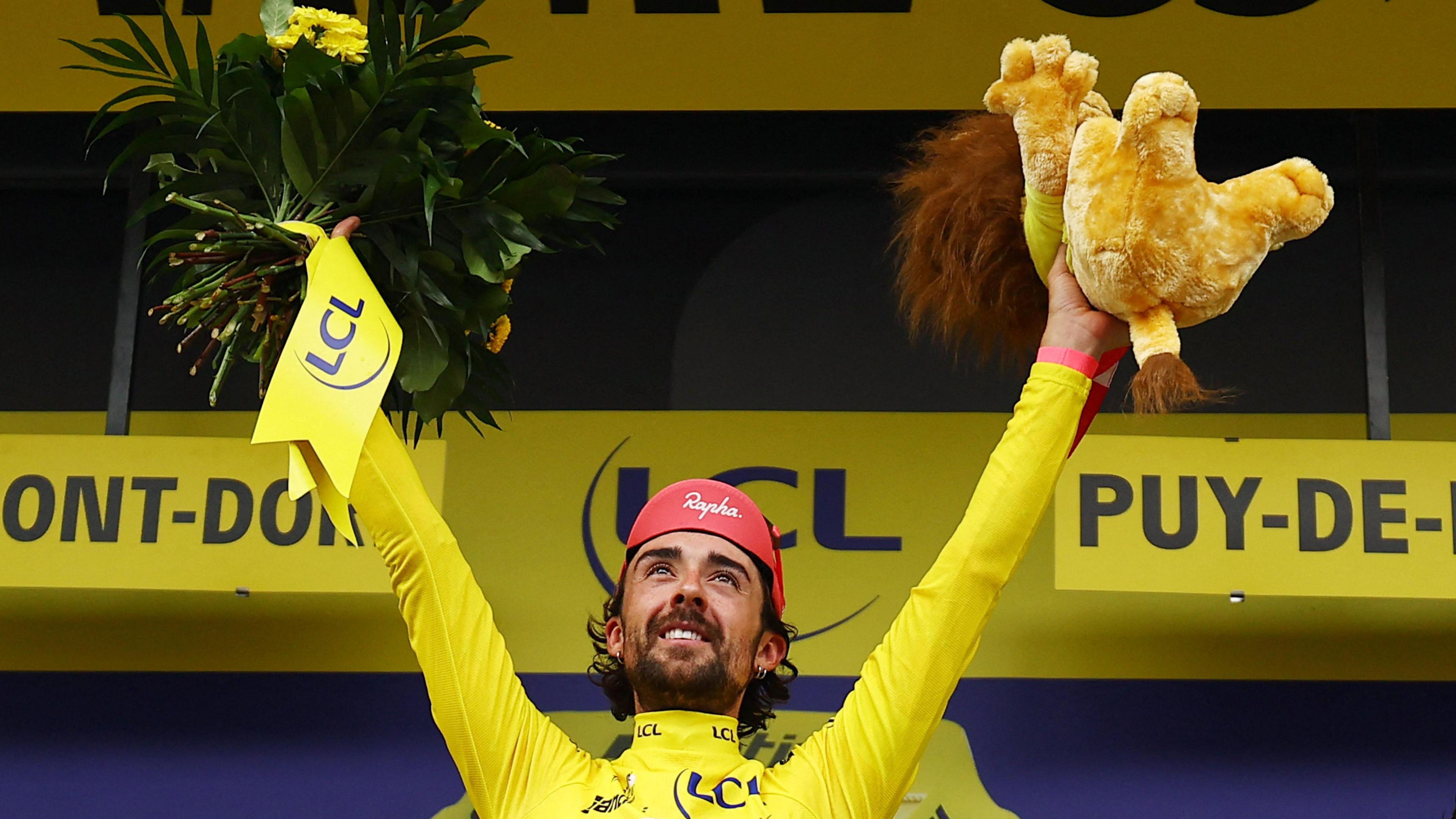 Ben Healy celebrates on the podium wearing the yellow jersey after stage six of the Tour De France. He is holding a stuffed animal in his left hand and flowers in his right.
