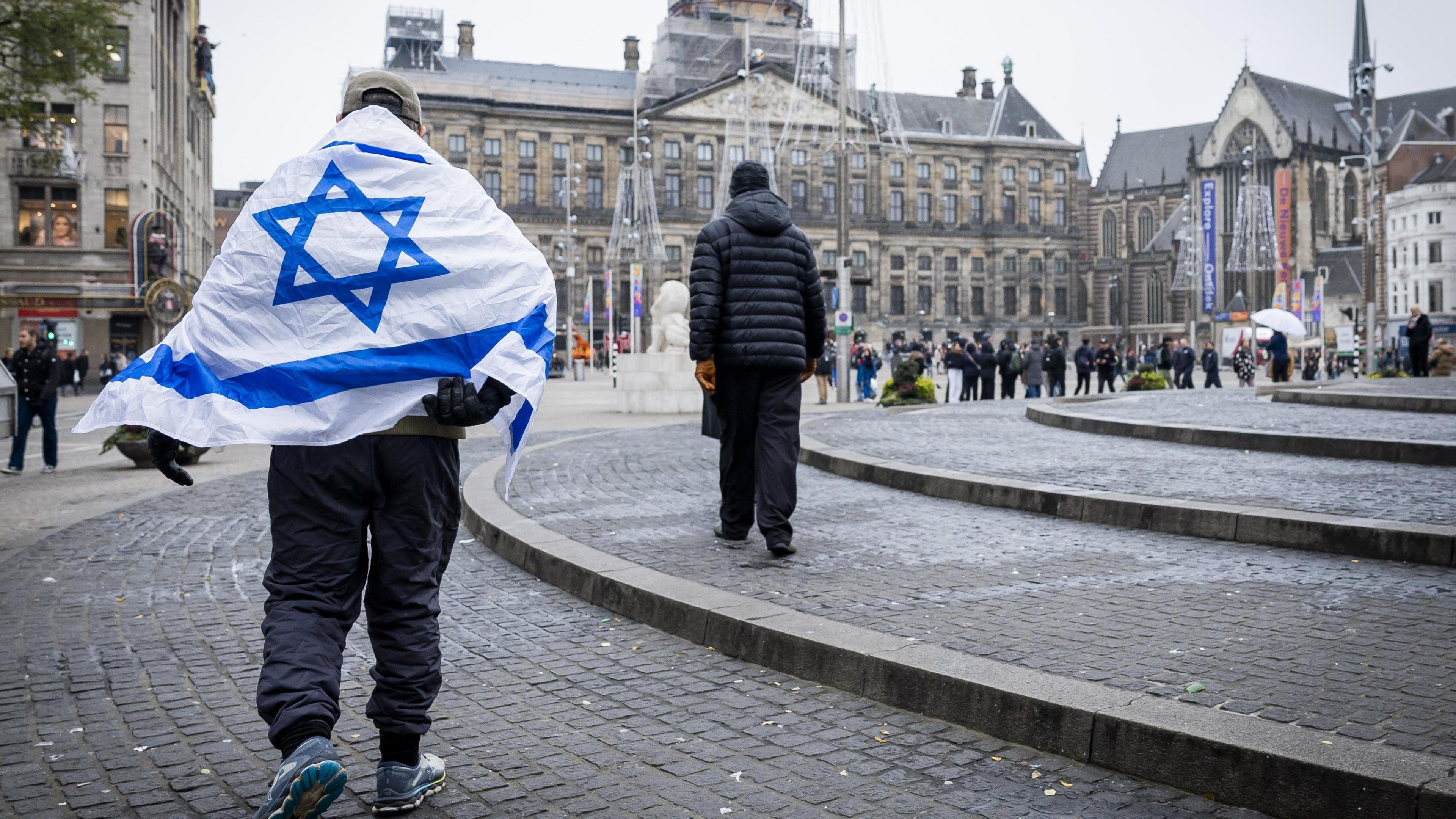A man wearing a blue and white Israeli flag on his shoulders walks around Dam Square in Amsterdam, Netherlands. Tall historic buildings can be seen in the background and people are standing around in the distance.