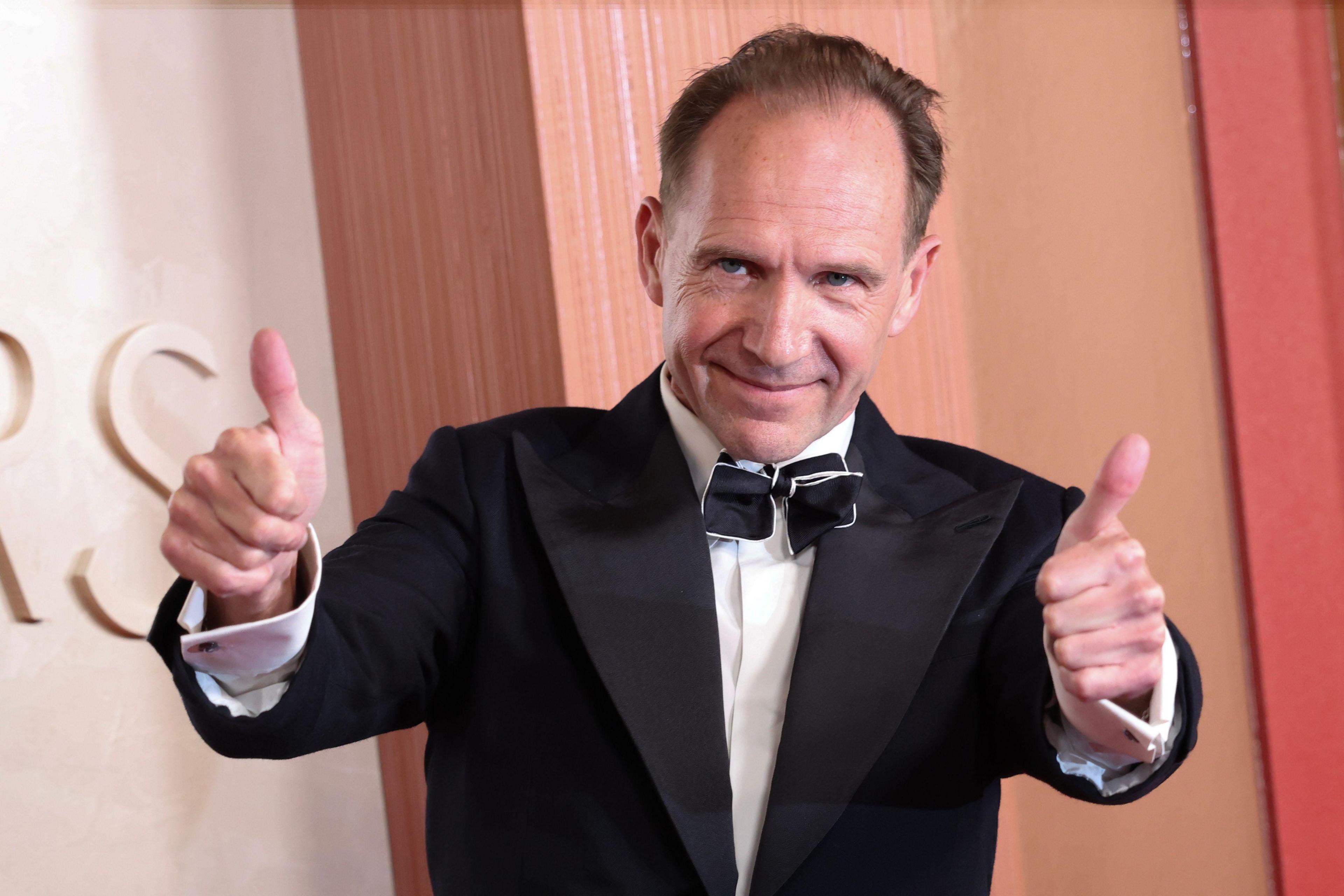 Ralph Fiennes on the red carpet. He is smiling and is holding both thumbs up.