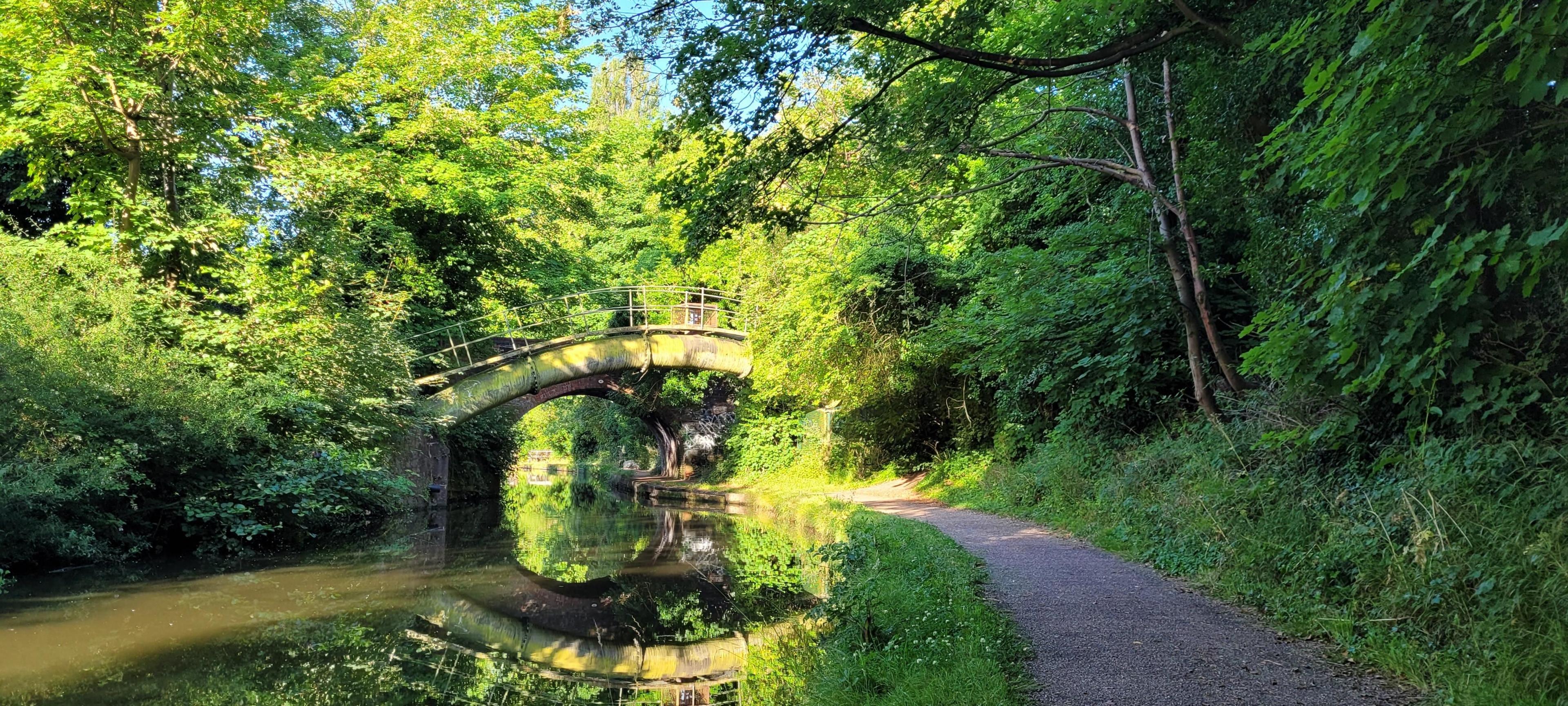 Canal and towpath in Hall Green, with sun shining through trees. A bridge over the canal is visible in the distance.