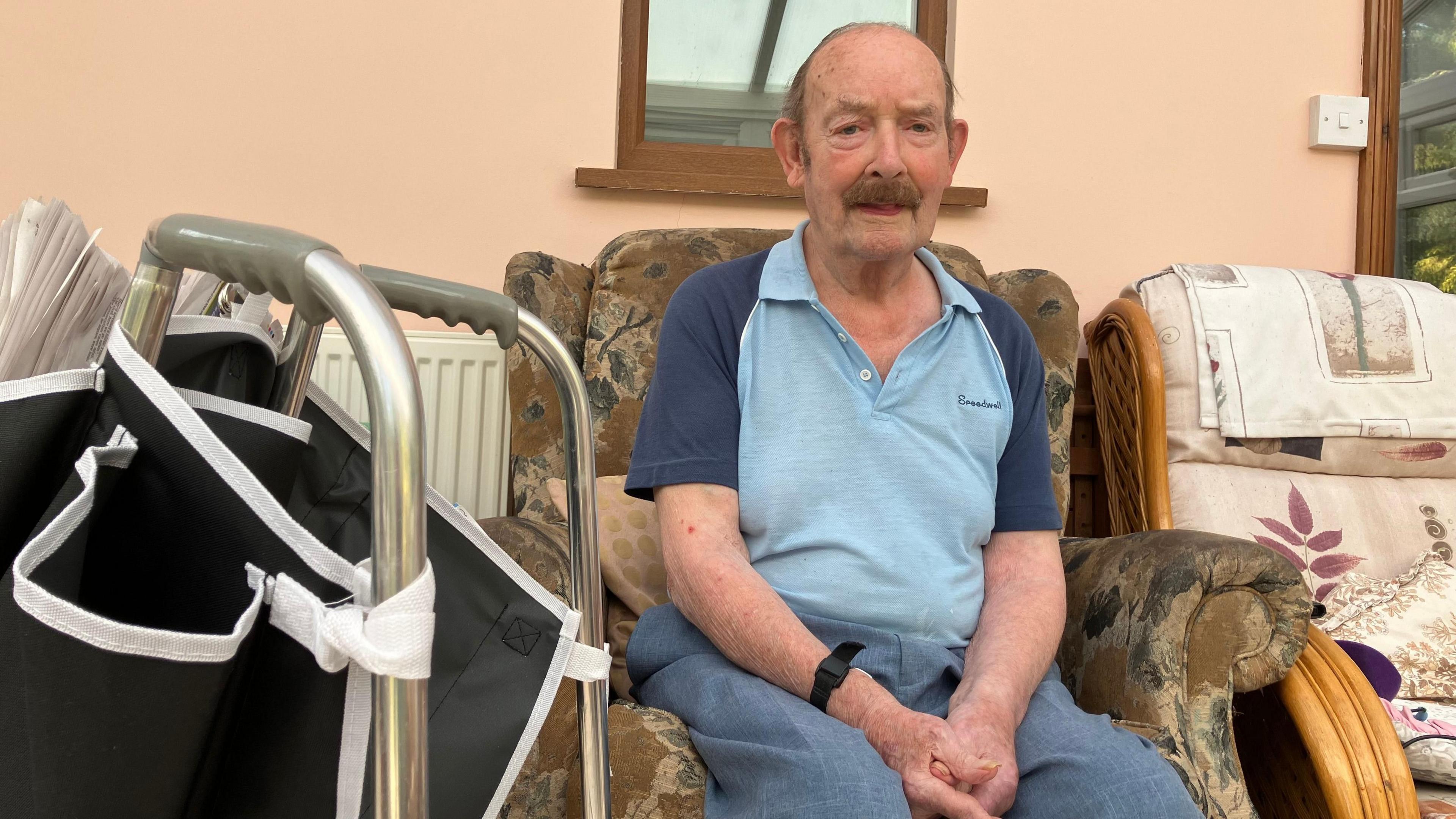 Bernard Woollaston is sitting on an armchair in his home, with a walking frame to his right. He is looking towards the camera, with his hands clasped together on his lap.