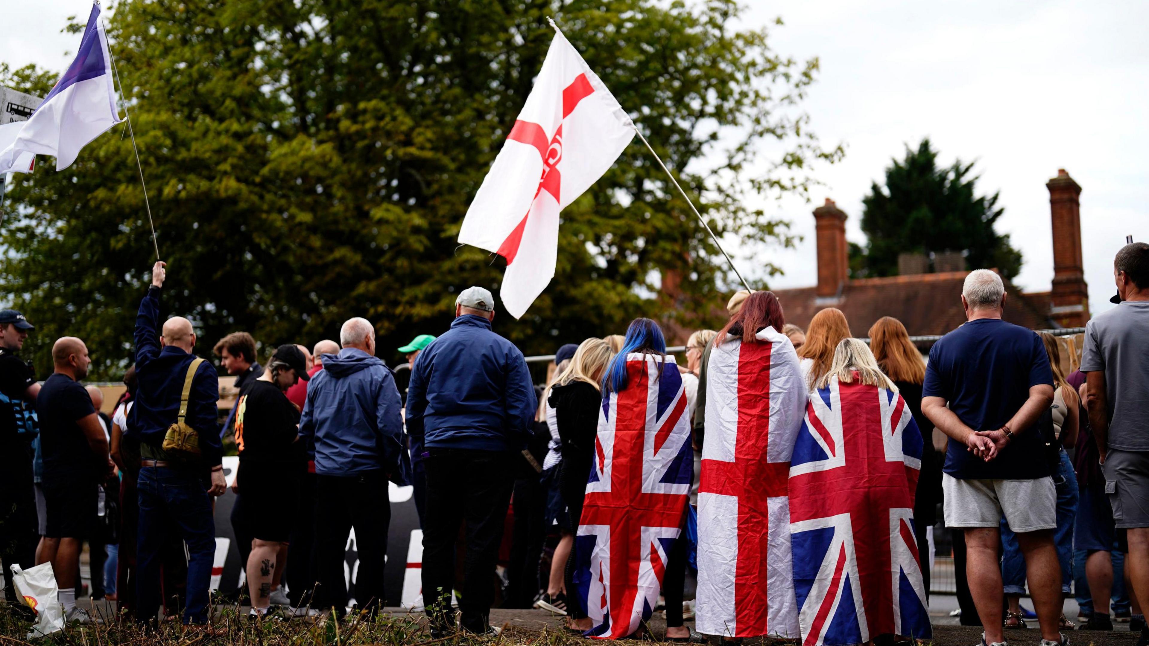 The backs of a crowd of people. Some are waving St George flags while others have the Union Jack flag wrapped around them.